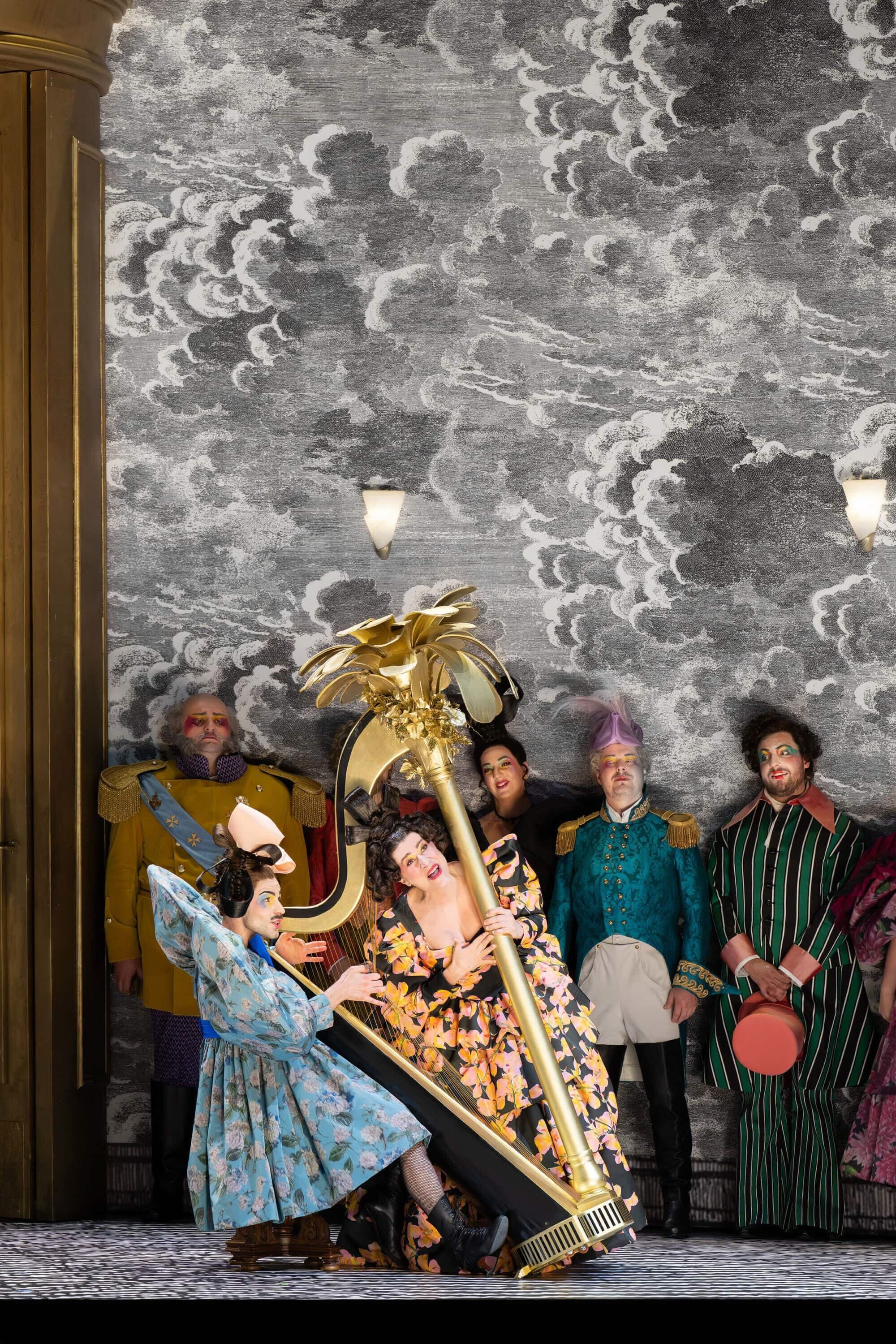 Mehrere Darsteller in bunten, extravaganten Kostümen posieren mit einer goldenen Harfe auf einer Bühne.