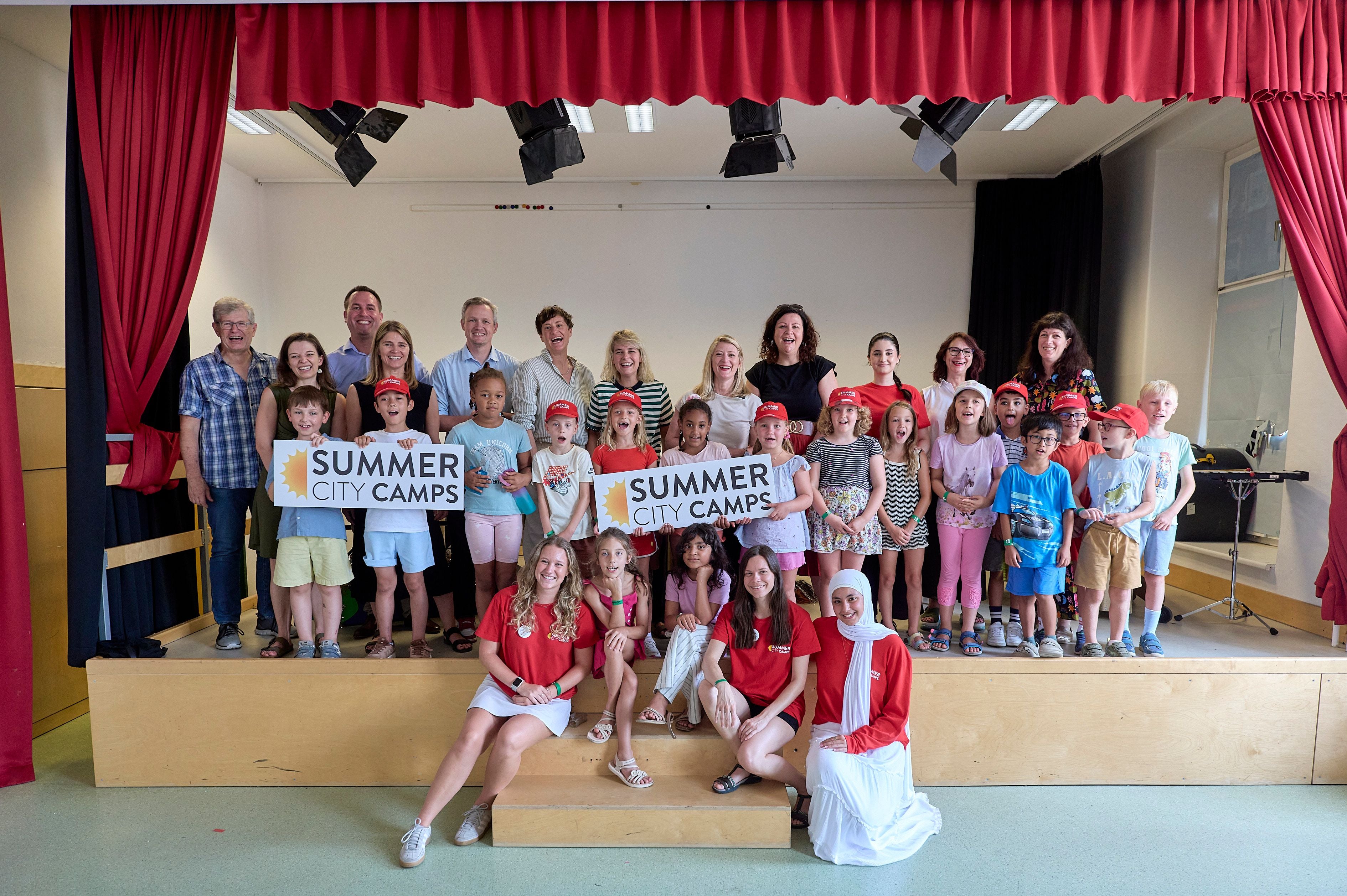 Eine Gruppe von Kindern und Erwachsenen posiert draußen mit einem „Summer City Camps“-Schild.