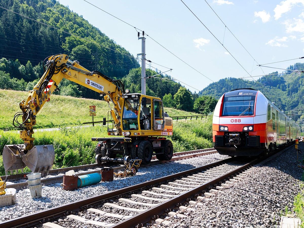 Ein Bagger und ein ÖBB-Zug stehen nebeneinander auf Gleisen in einer grünen Hügellandschaft.