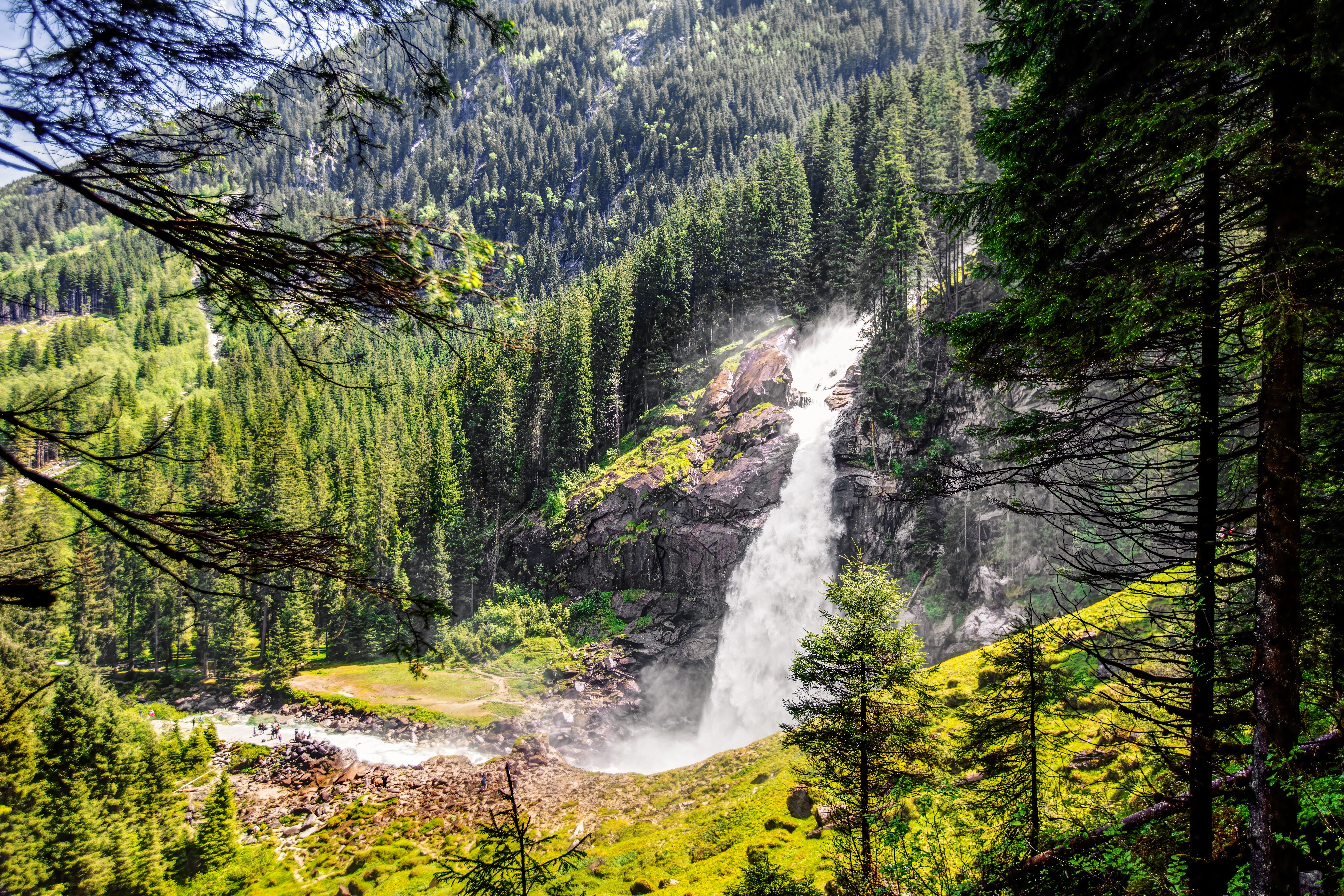 Mächtiger Wasserfall umgeben von dichten grünen Nadelwäldern in einer Gebirgslandschaft.