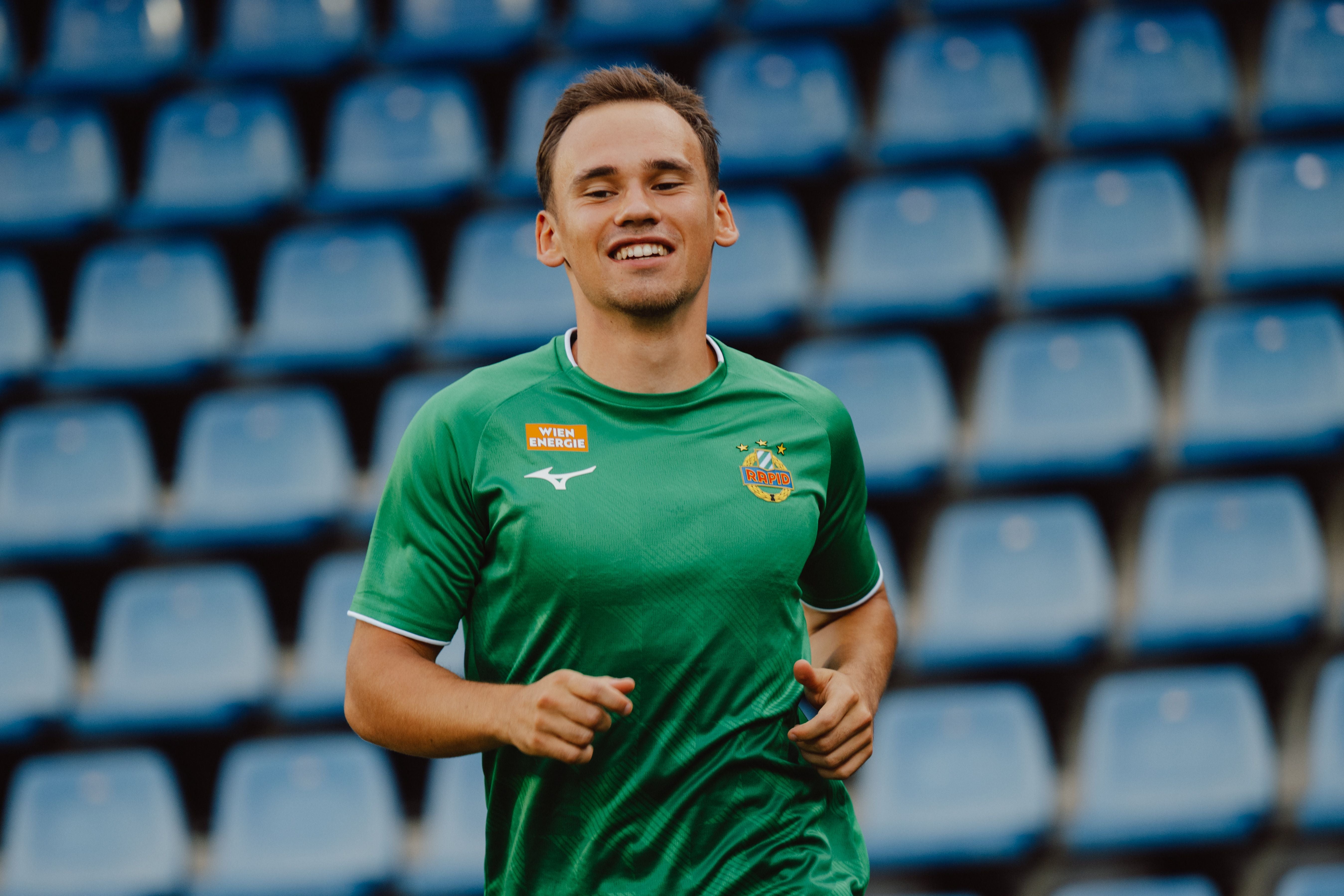 Ein Fußballspieler im grünen Trikot joggt lächelnd vor leeren blauen Stadionrängen.