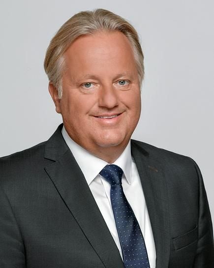 Ein Mann im Anzug lächelt vor neutralem Hintergrund in die Kamera.
