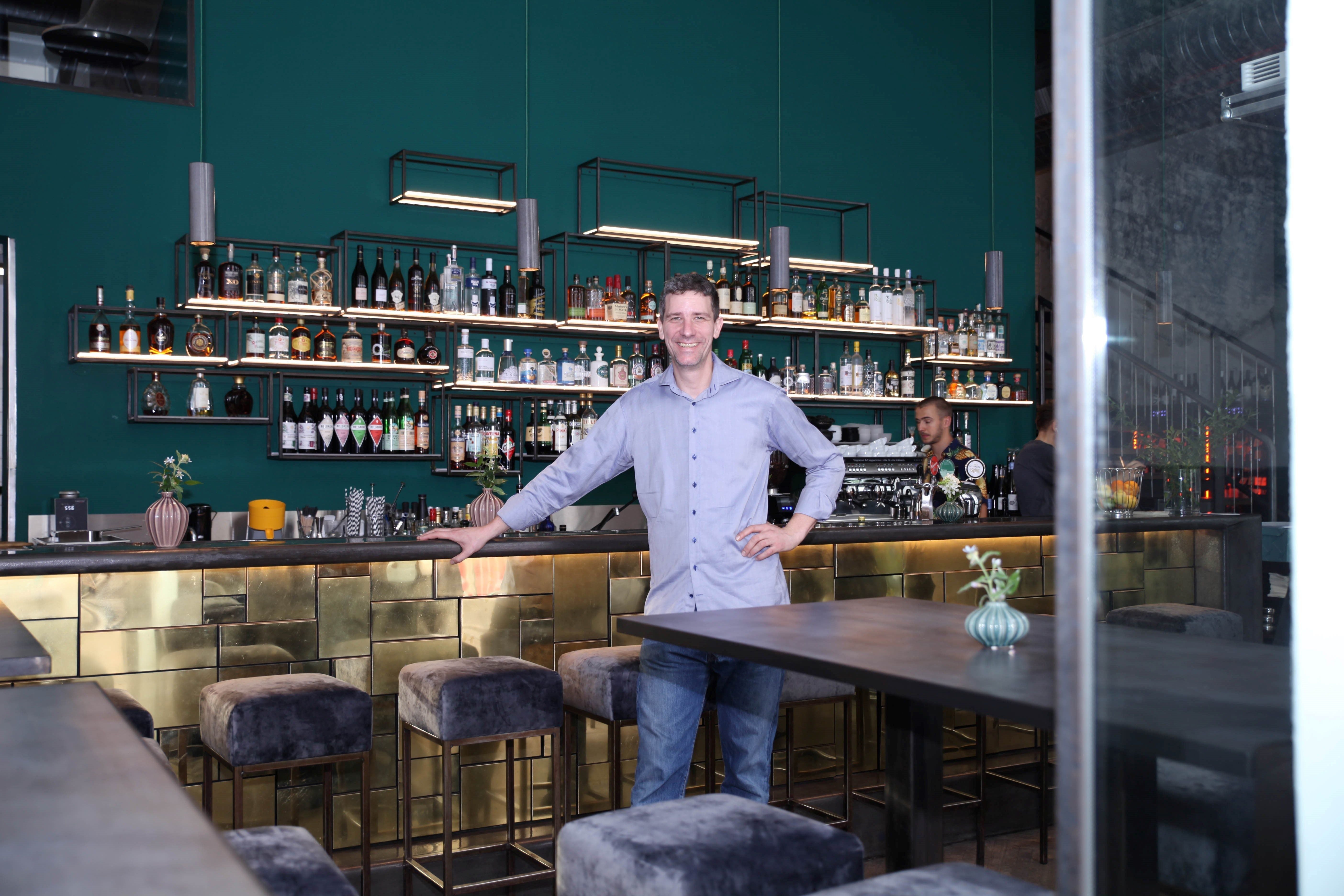 Mann steht lächelnd an einer eleganten Bar mit goldener Front und Flaschenregalen im Hintergrund.