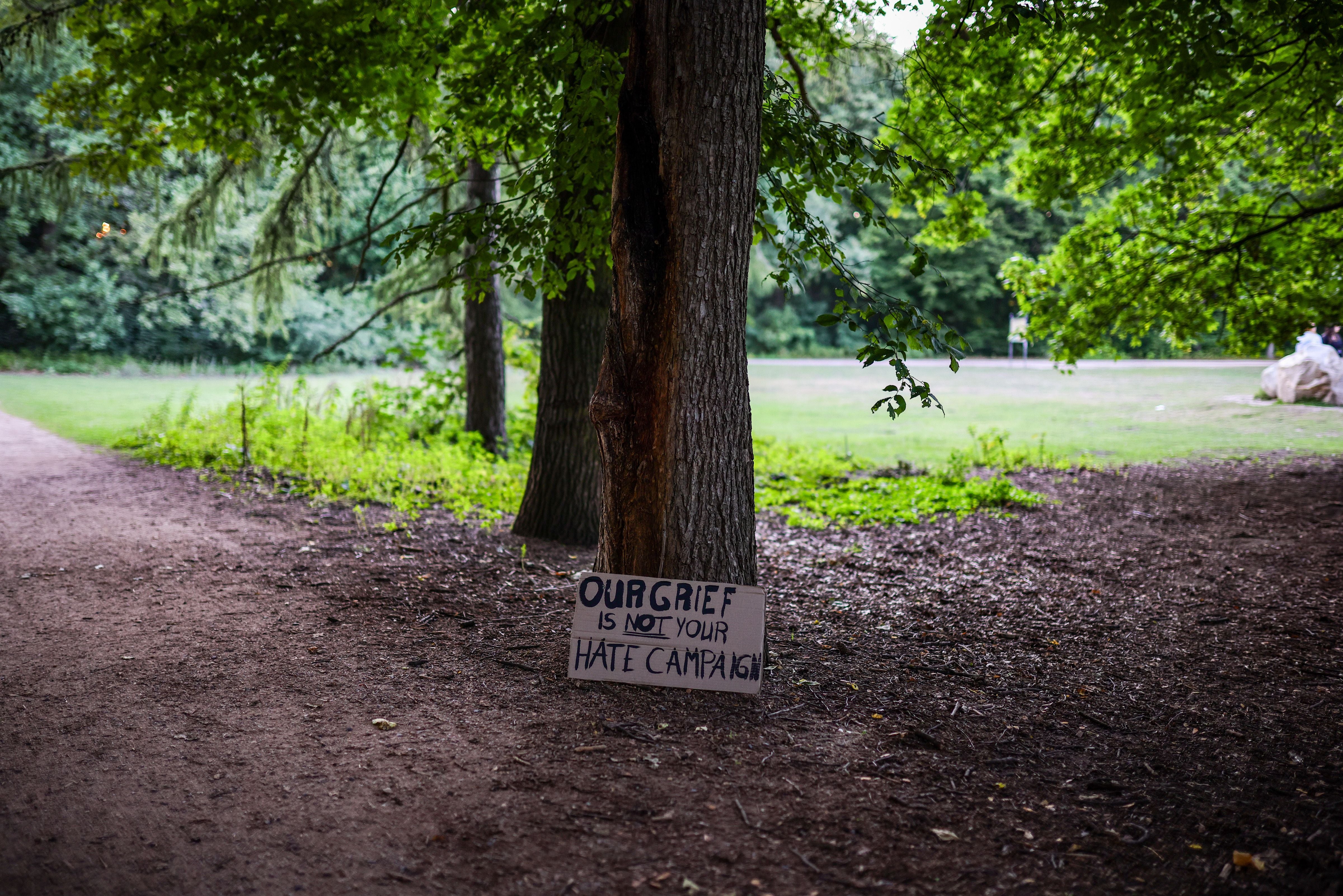 Ein Pappschild mit der Aufschrift „Our grief is NOT your hate campaign“ lehnt an einem Baum im Park.