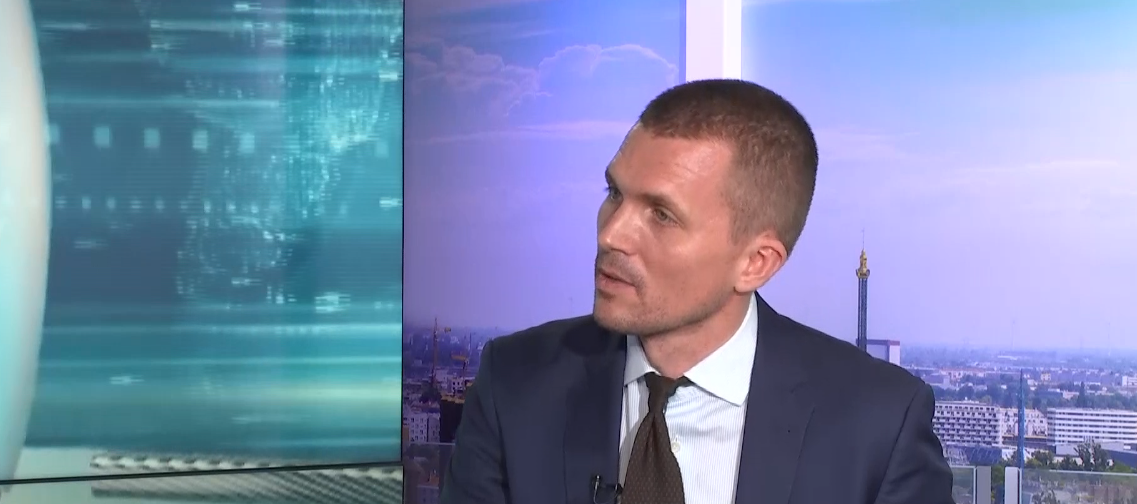 Ein Mann im Anzug wird in einem Fernsehstudio interviewt, im Hintergrund eine Stadtansicht.