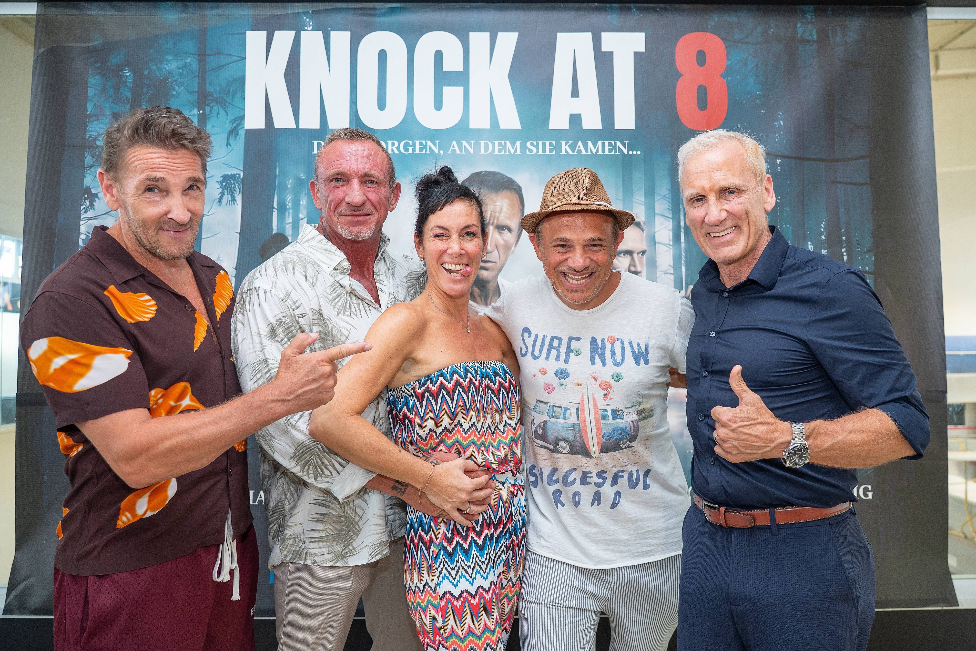 Fünf fröhliche Menschen posieren vor einem Filmplakat von „Knock at 8“.