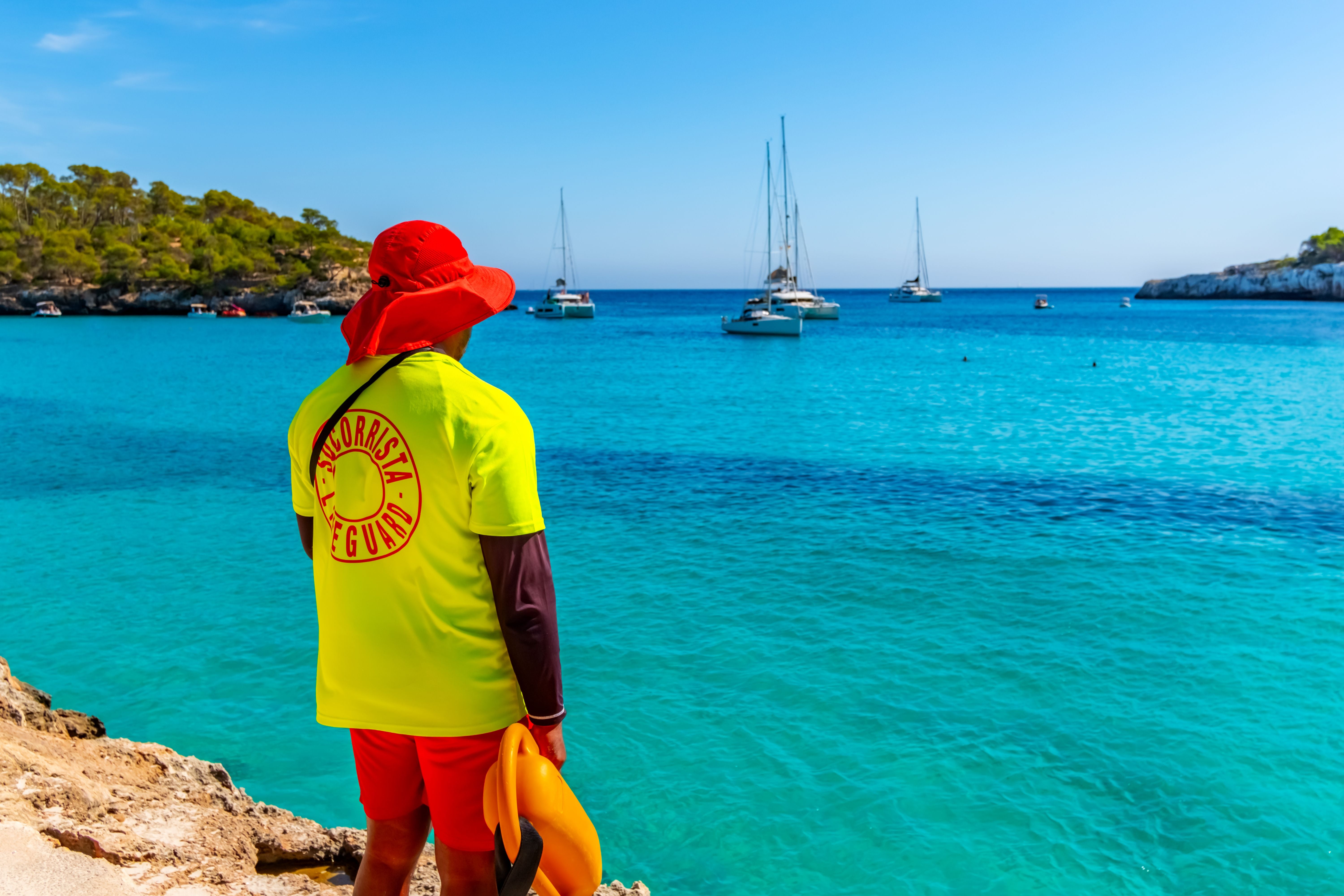 Rettungsschwimmer mit rotem Hut am Strand von Cala Mondrago, türkisblaues Meer und Boote im Hintergrund.