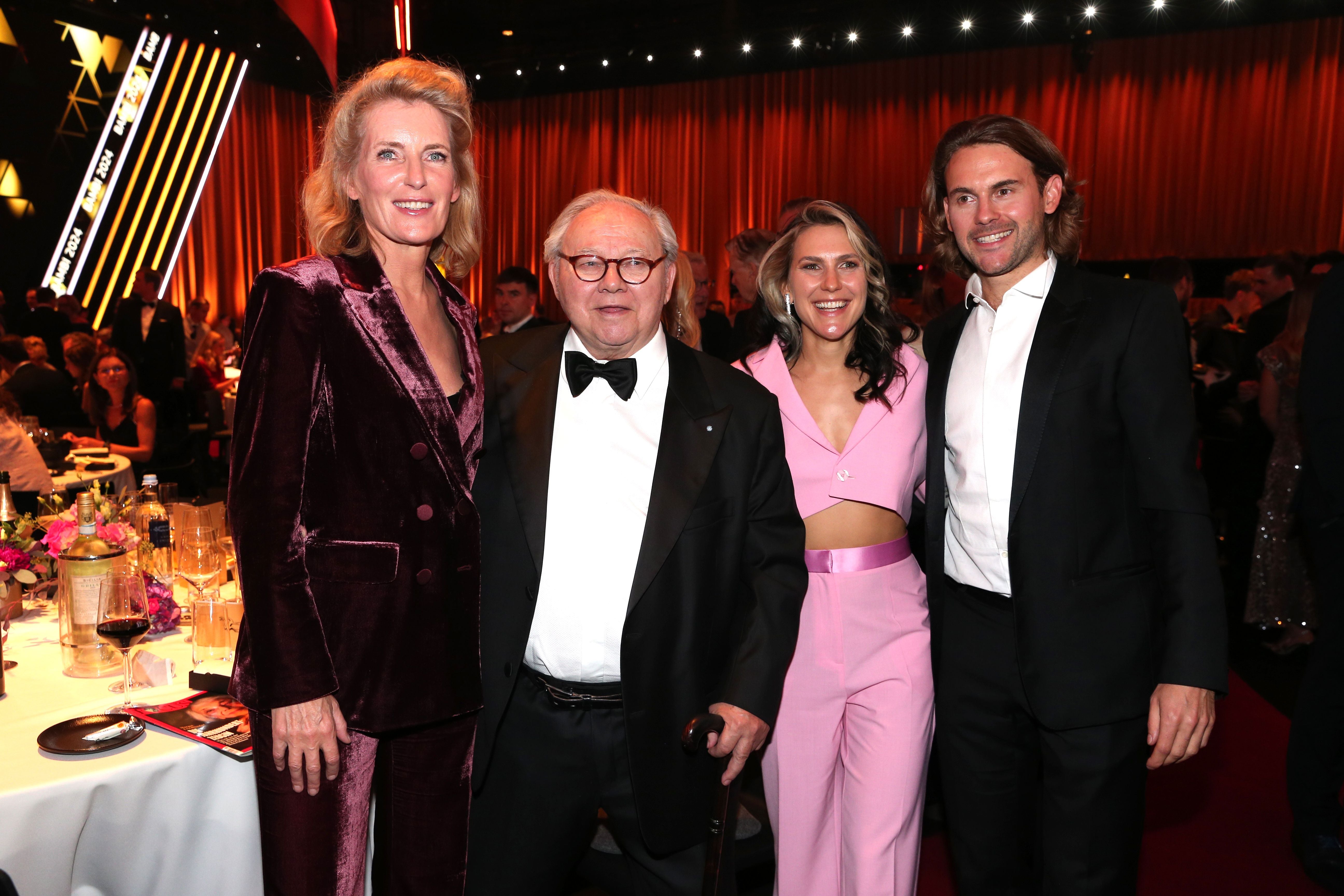Vier elegant gekleidete Personen posieren gemeinsam bei einer Gala mit festlich beleuchtetem Hintergrund.