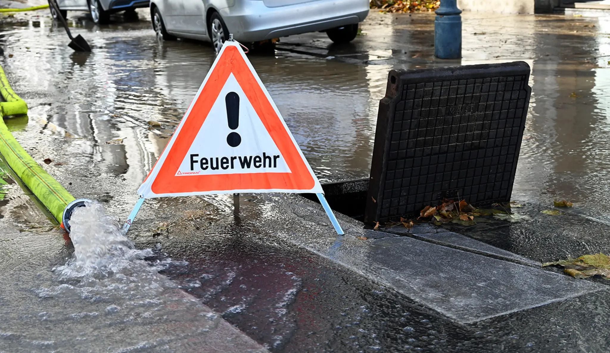 Dreieckiges Warnschild "Feuerwehr" bei Wasserrohrbruch auf nasser Straße mit offenem Gully.