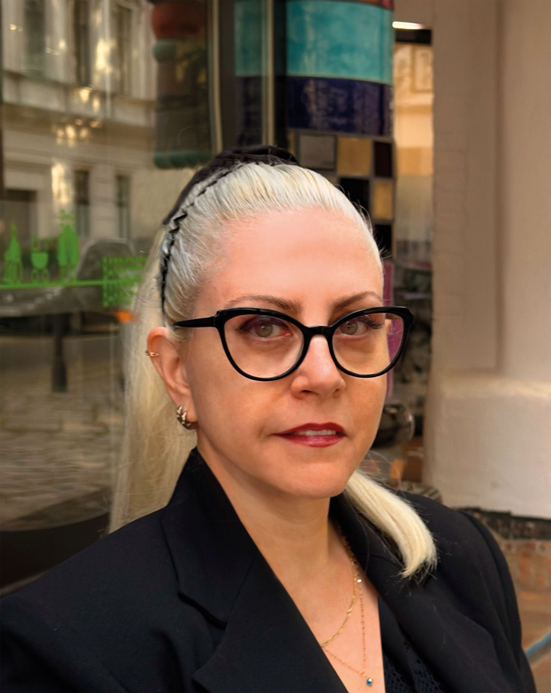 Frau mit blonden Haaren, schwarzer Brille und schwarzem Blazer steht draußen vor einem Gebäude.