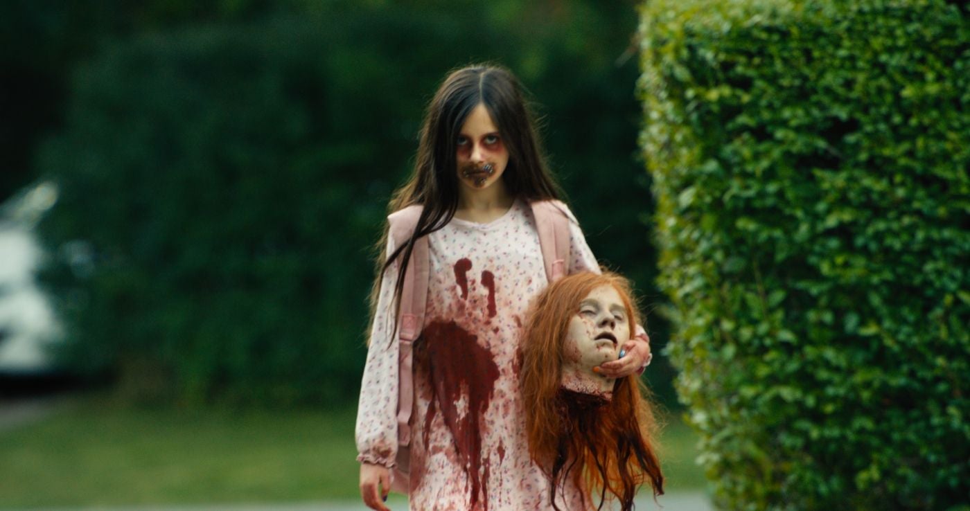 Mädchen im blutigen Nachthemd hält abgetrennten Puppenkopf, Horror-Make-up im Gesicht.