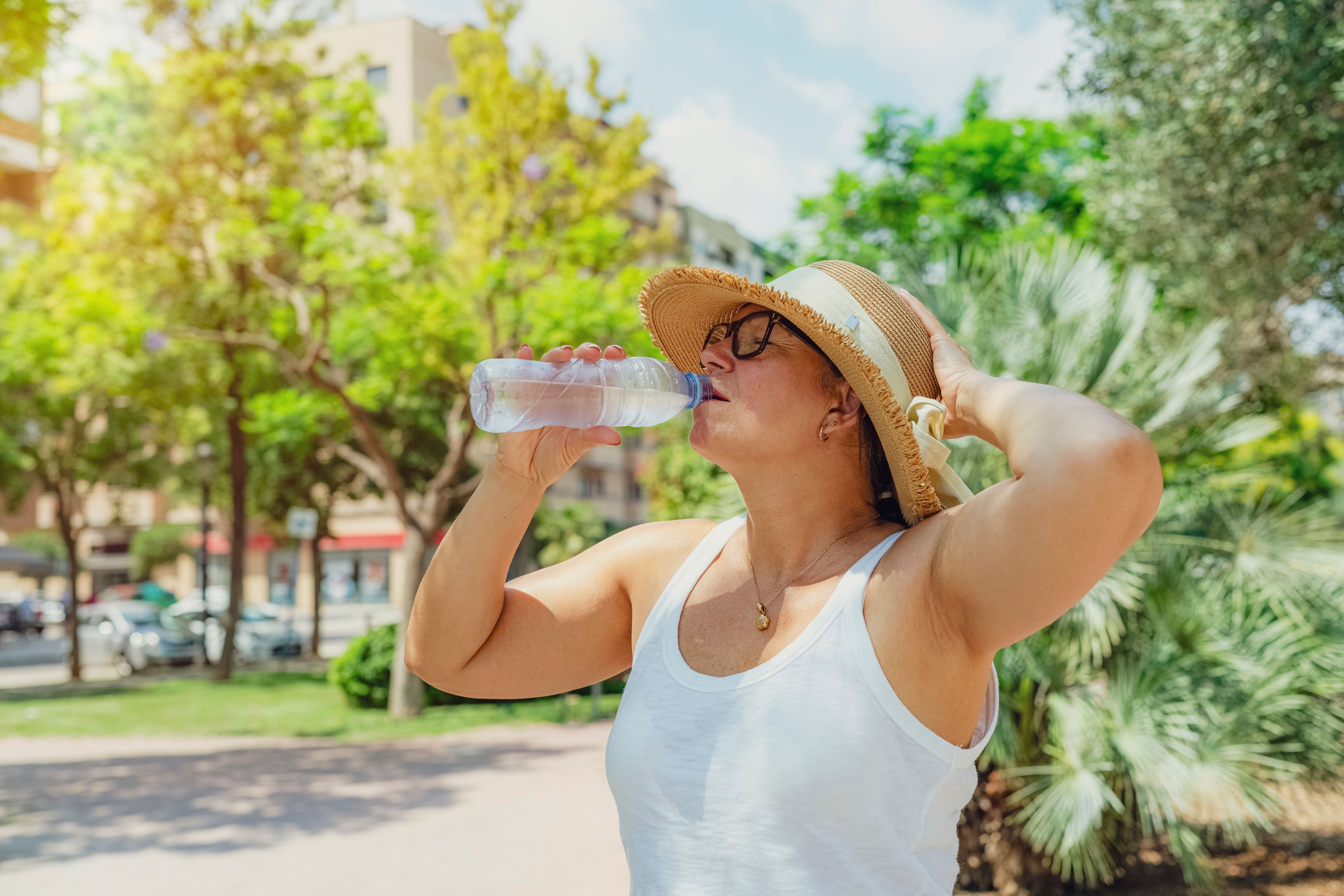 Frau mit Sonnenhut trinkt im Park Wasser und schützt sich vor der Sommerhitze.