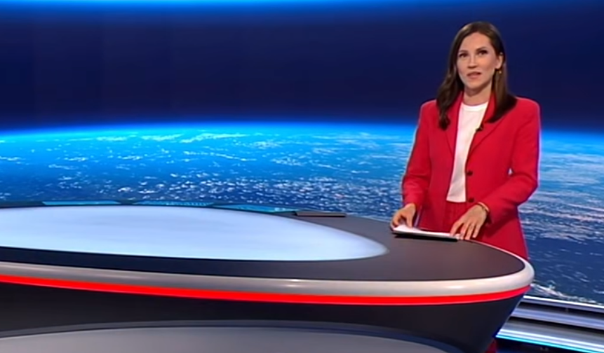 Nachrichtensprecherin im roten Anzug steht in einem Fernsehstudio mit Weltall-Hintergrund.