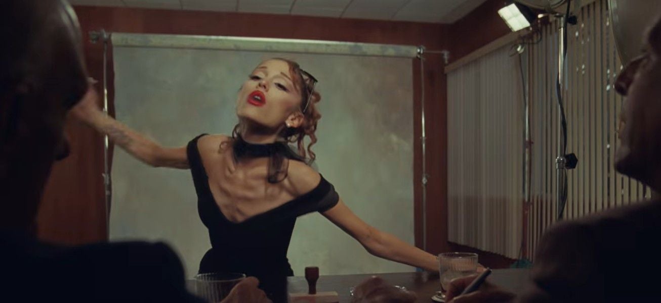 Ariana Grande posiert in einem Retro-Fotostudio vor zwei Personen mit Notizblock und Getränk.