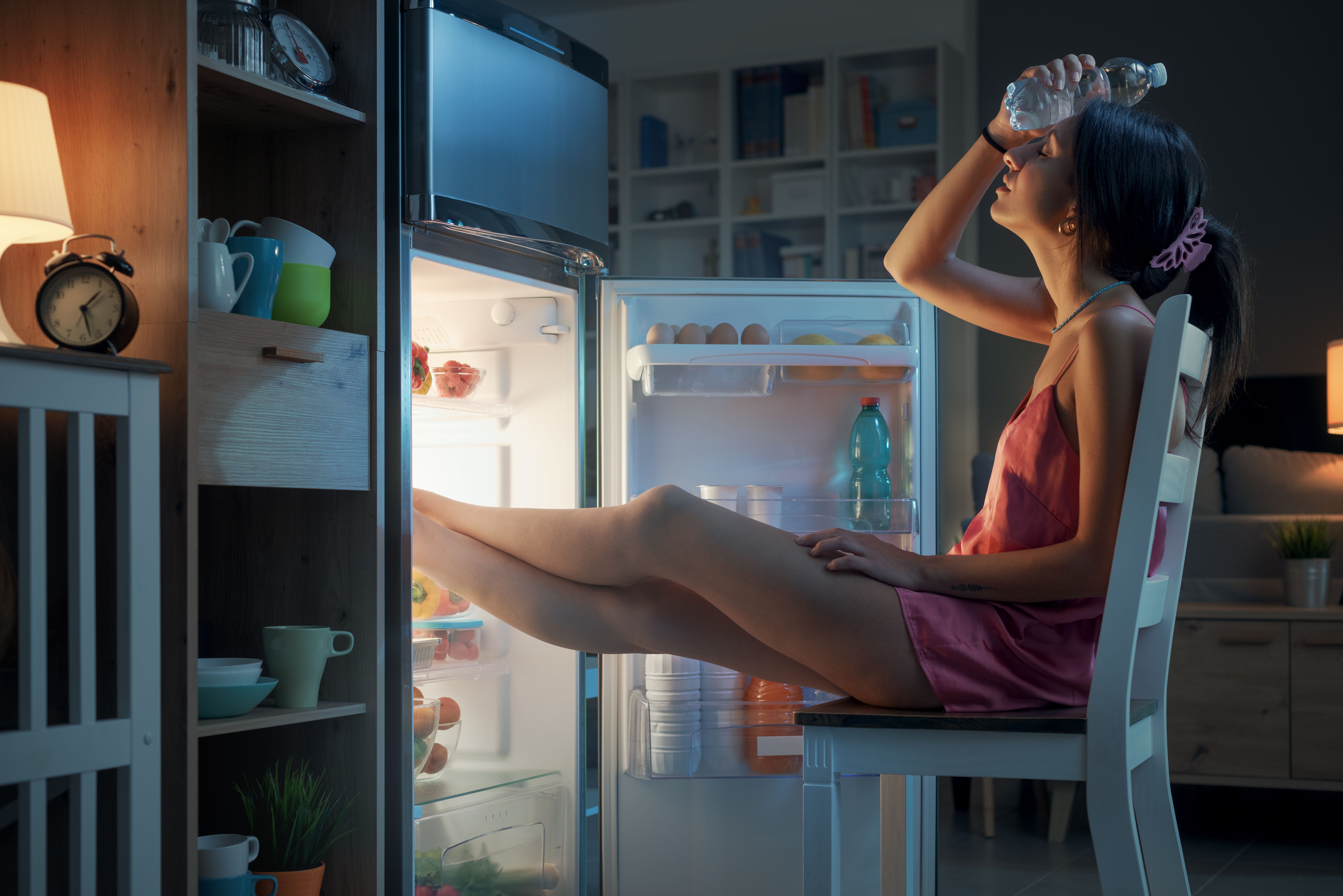 Frau sitzt nachts mit den Füßen im Kühlschrank und kühlt sich mit einer Wasserflasche ab.