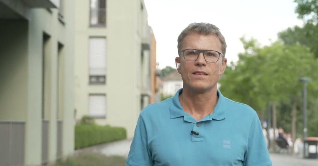 Ein Mann mit Brille und blauem Polohemd steht draußen vor Wohngebäuden und Bäumen.