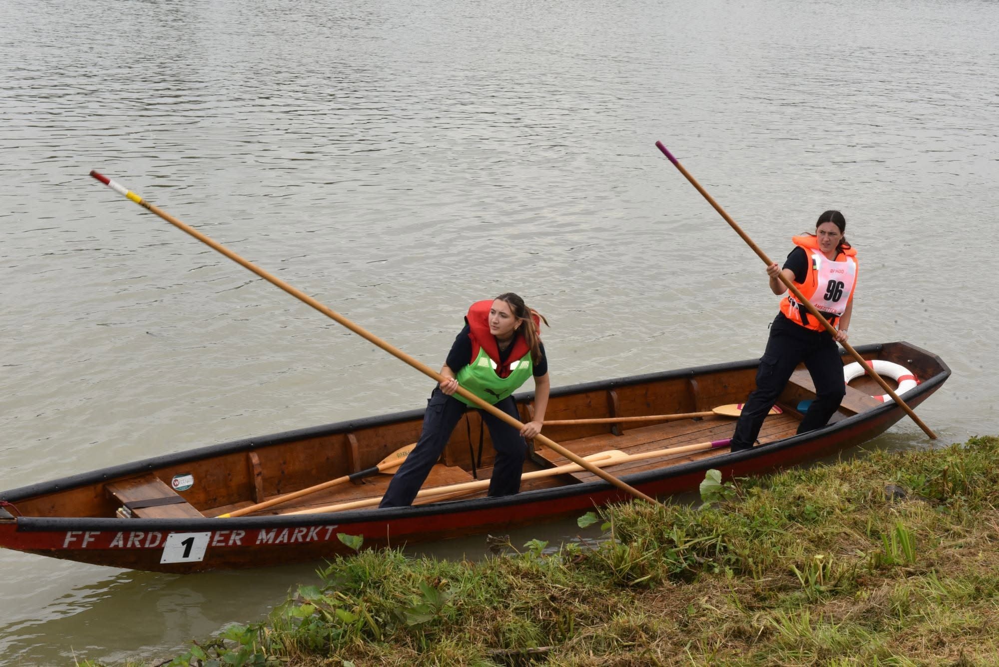 Zwei Frauen rudern mit Stangen ein Boot nah am Ufer eines Flusses entlang.