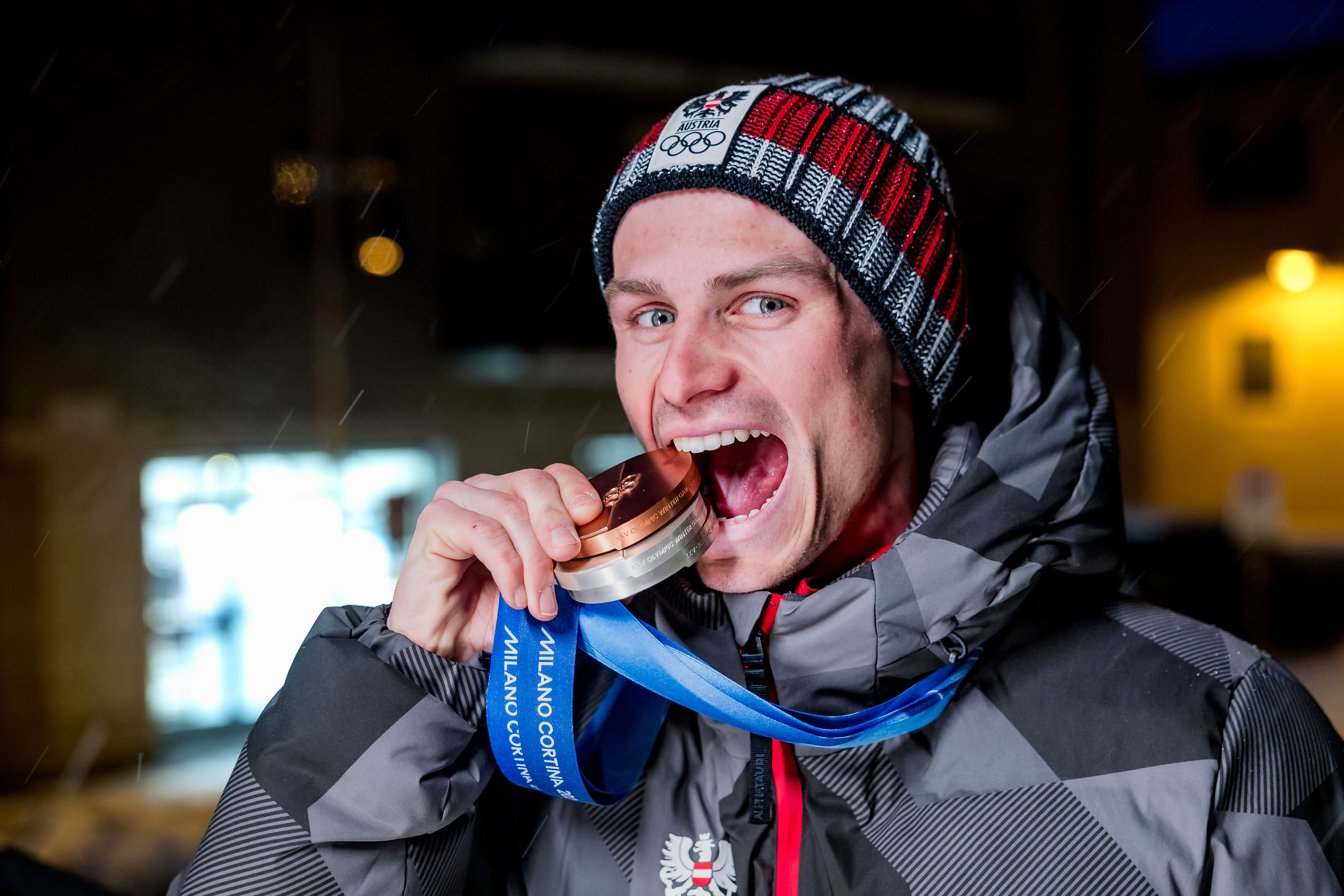 Ein junger Mann beißt auf eine Medaille und trägt eine Winterjacke sowie eine Mütze mit Österreich-Logo.