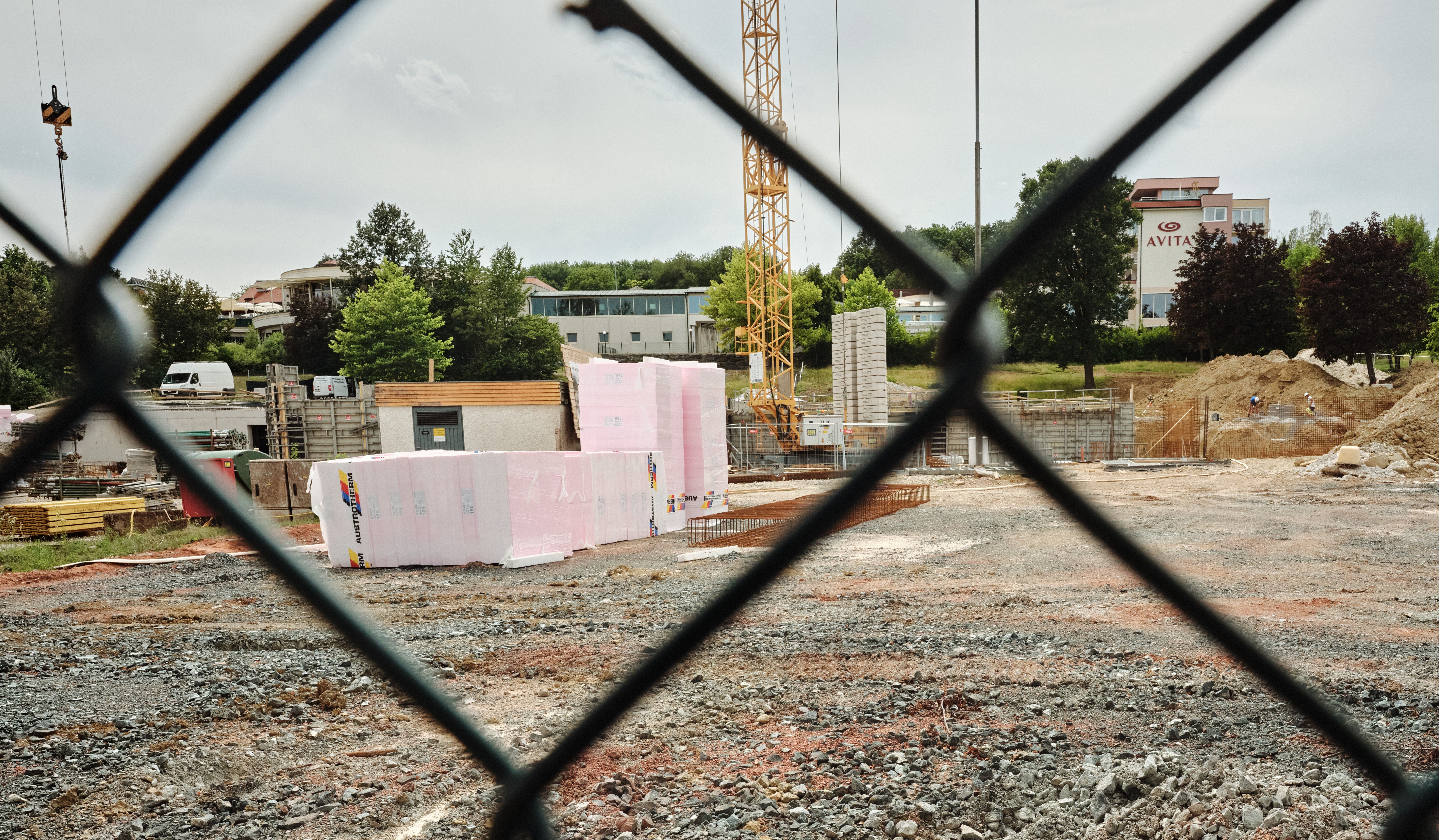 Blick durch einen Zaun auf eine Baustelle mit Baumaterial und Kran, im Hintergrund Gebäude.