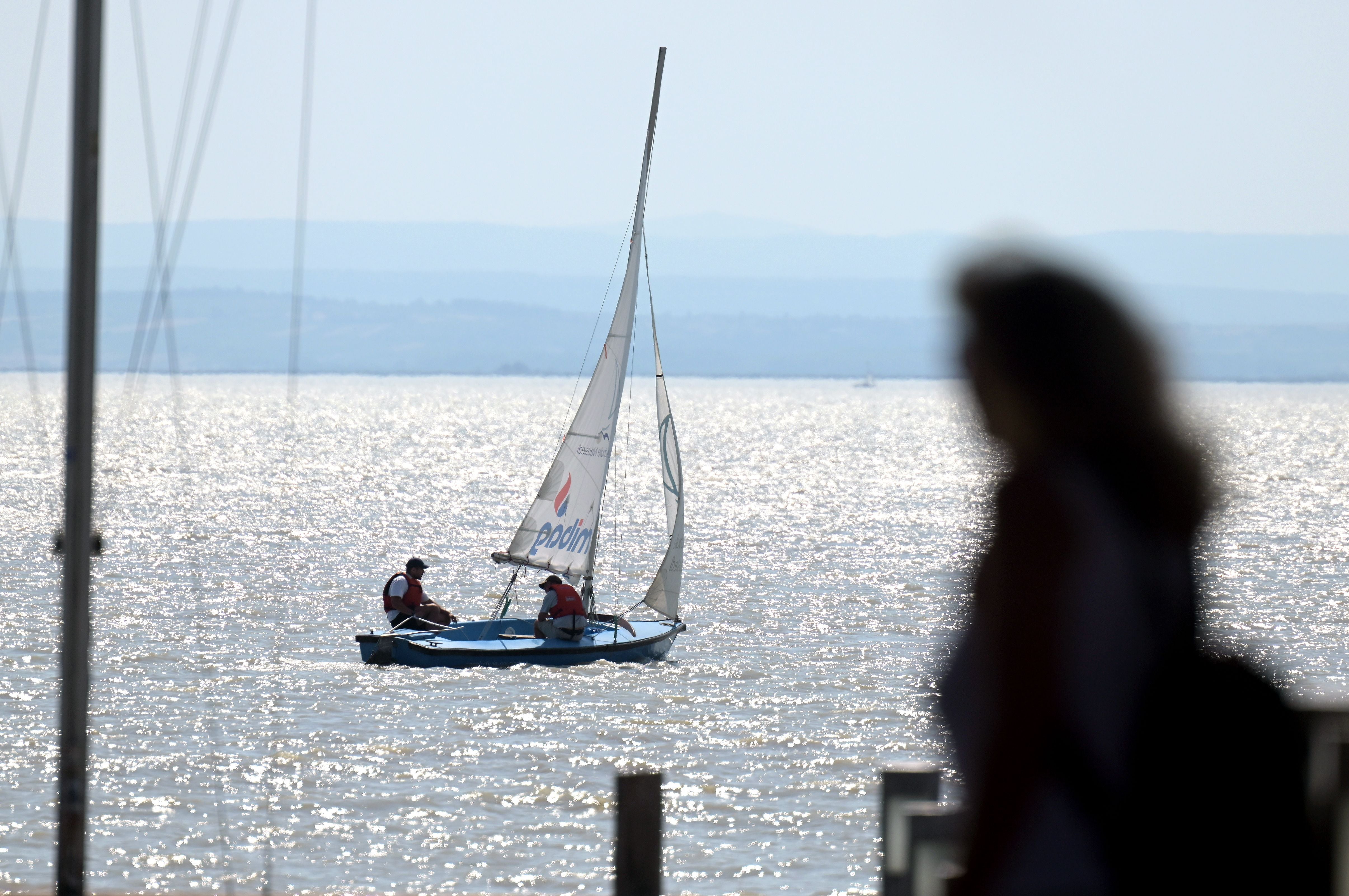 Segelboot mit zwei Personen auf dem glitzernden Neusiedler See, im Vordergrund eine unscharfe Person.