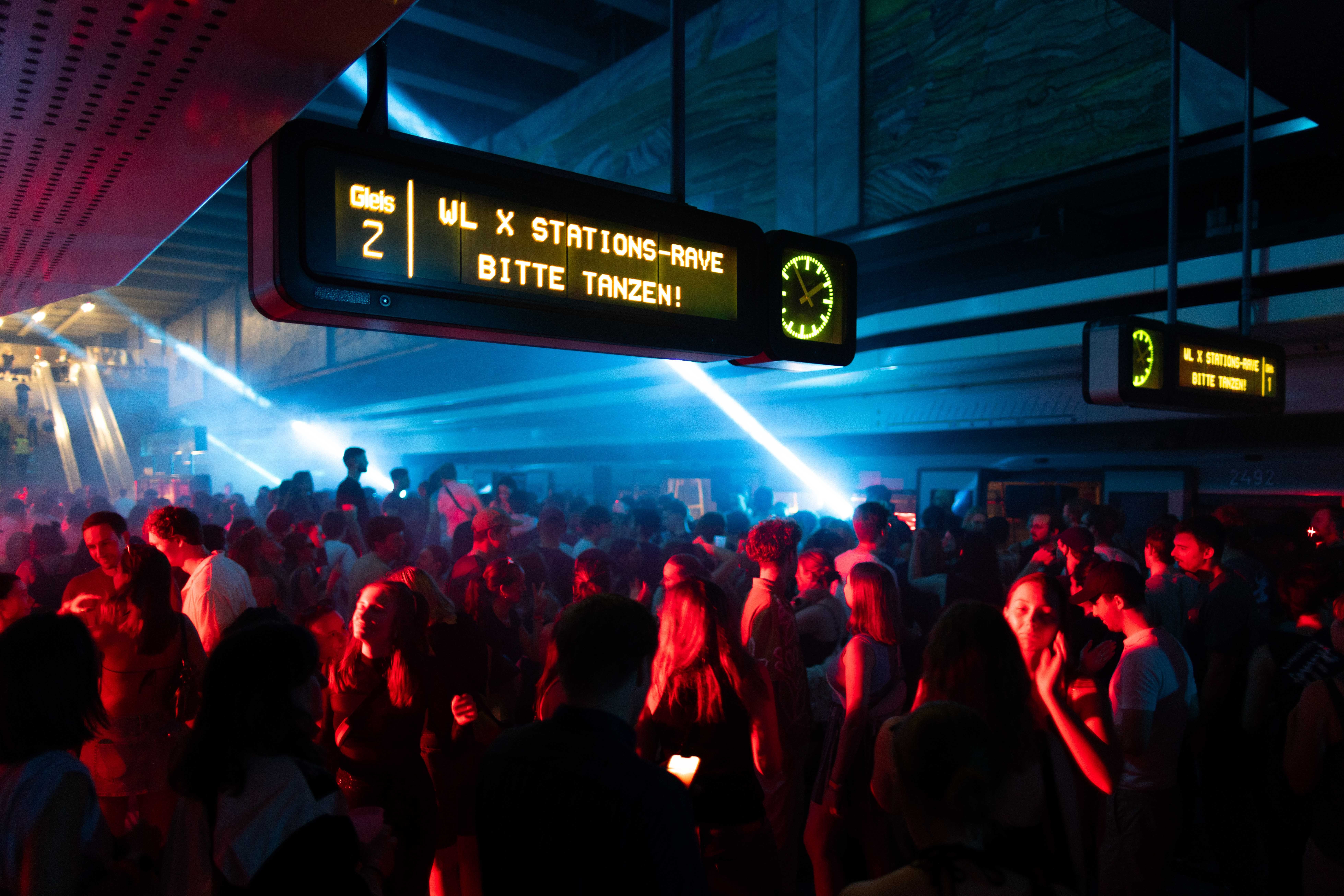 Viele junge Menschen tanzen bei einer Rave-Party in einer bunt beleuchteten U-Bahn-Station.
