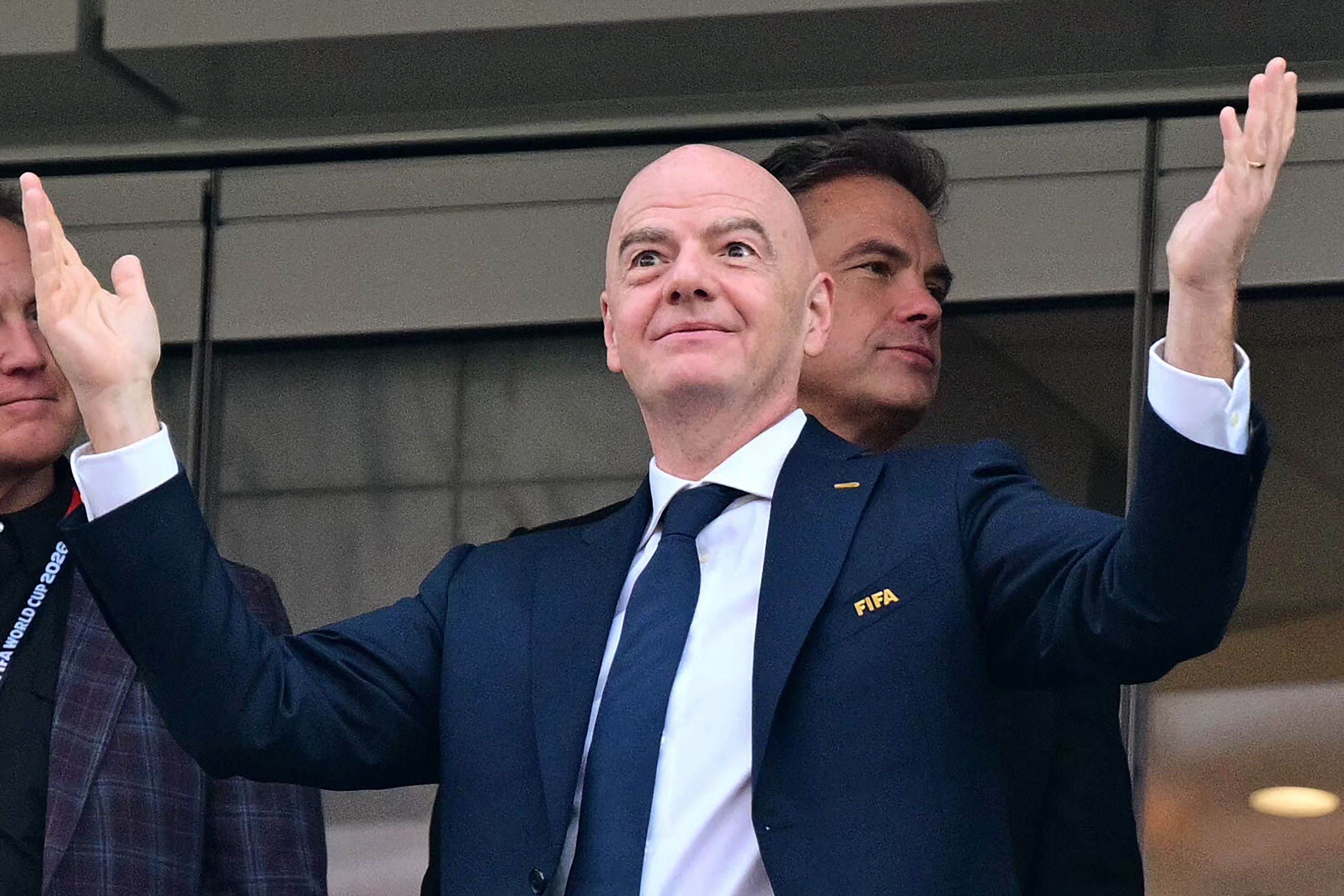 Ein Mann im Anzug mit erhobenen Armen steht in einem Stadion und trägt ein FIFA-Logo am Revers.
