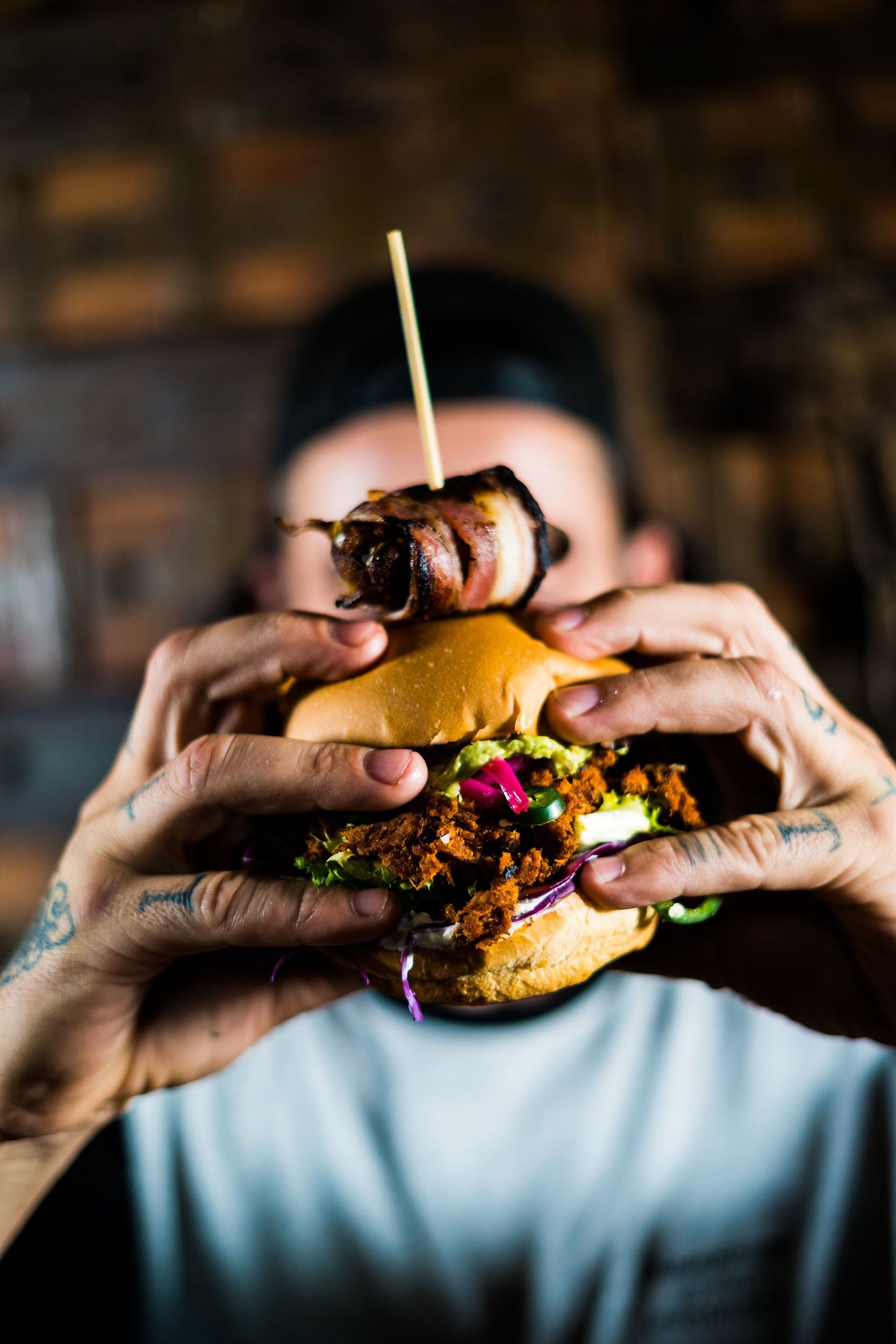 Person hält einen großen Burger mit verschiedenen Zutaten und Bacon in beiden Händen.