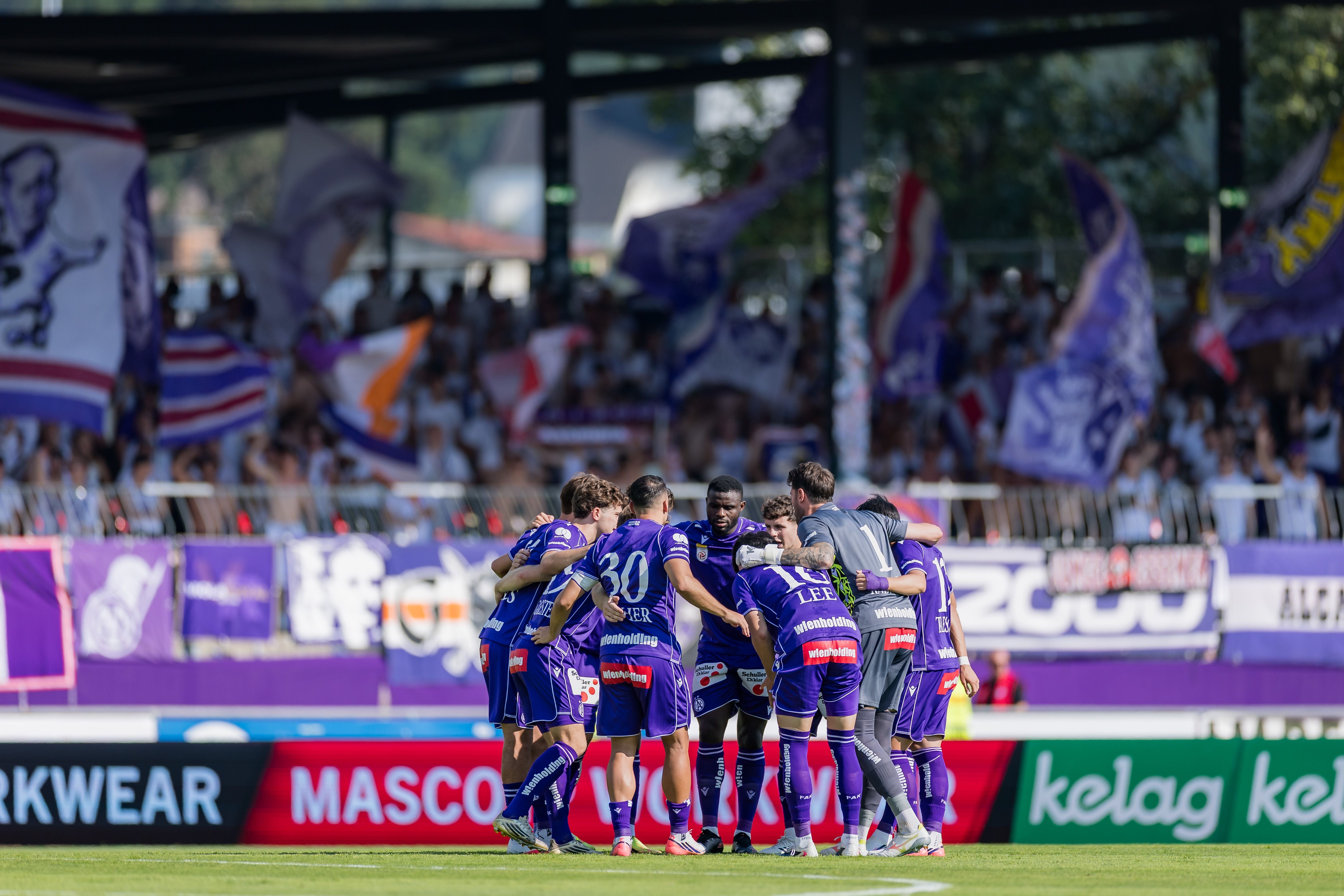 Fußballspieler von Austria Wien stehen im Kreis auf dem Spielfeld, Fans und Fahnen im Hintergrund.