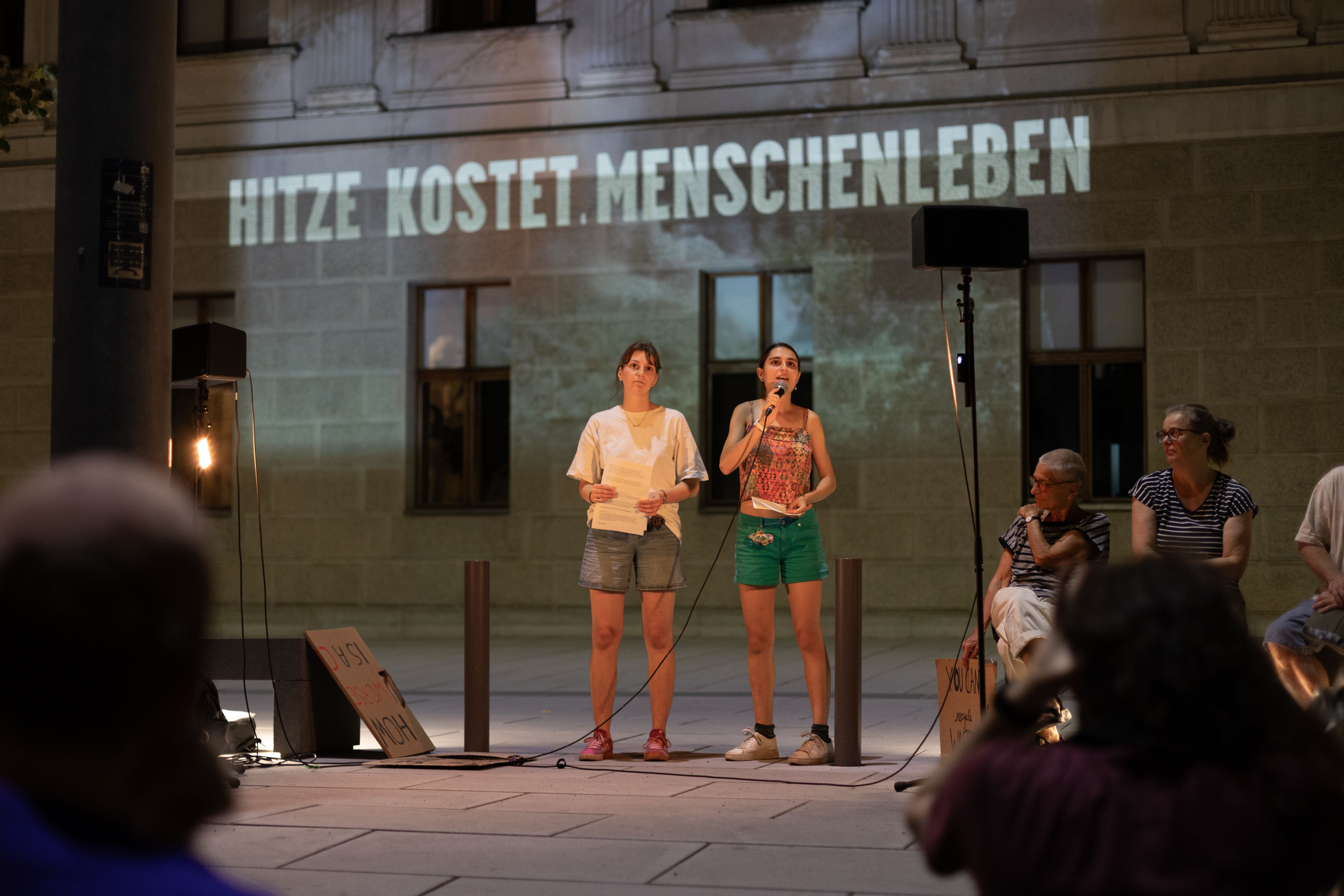 Zwei Frauen sprechen bei einer Hitze-Demo, im Hintergrund steht „Hitze kostet Menschenleben“.