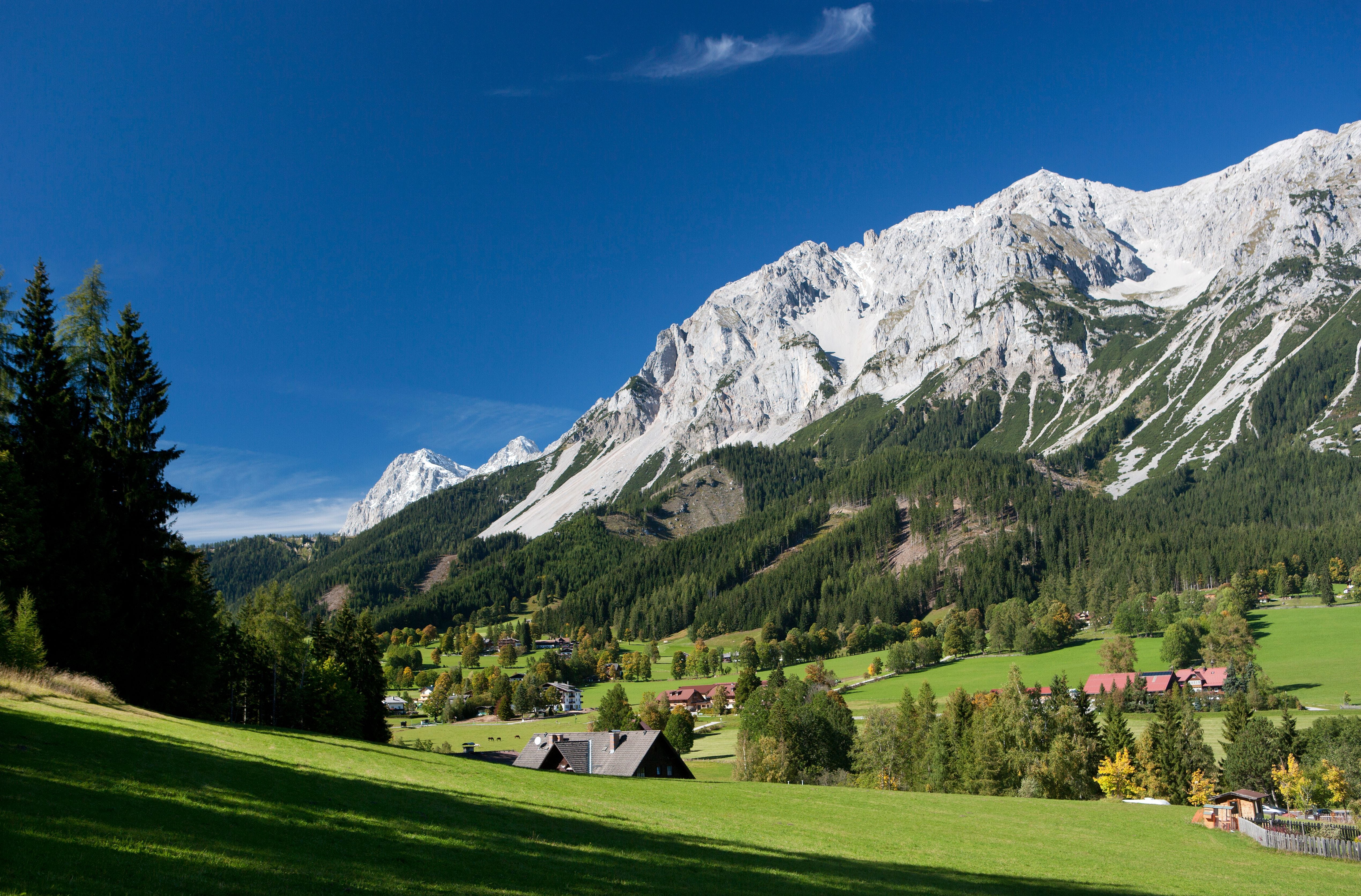 Blick auf ein grünes Tal mit Wiesen, Dörfern und schneebedeckten Bergen im Hintergrund.