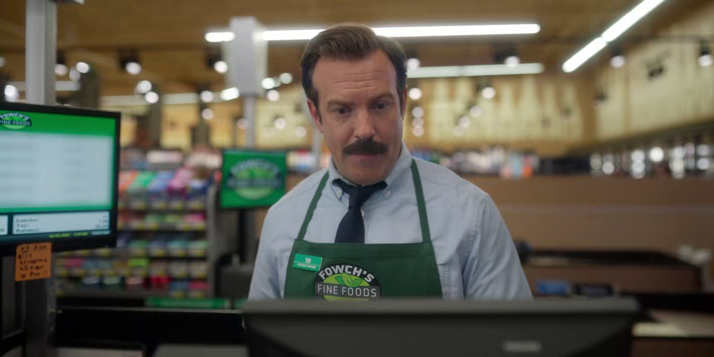 Mann mit Schnurrbart und grünem "Fowch's Fine Foods"-Schürze steht an einer Supermarktkasse.
