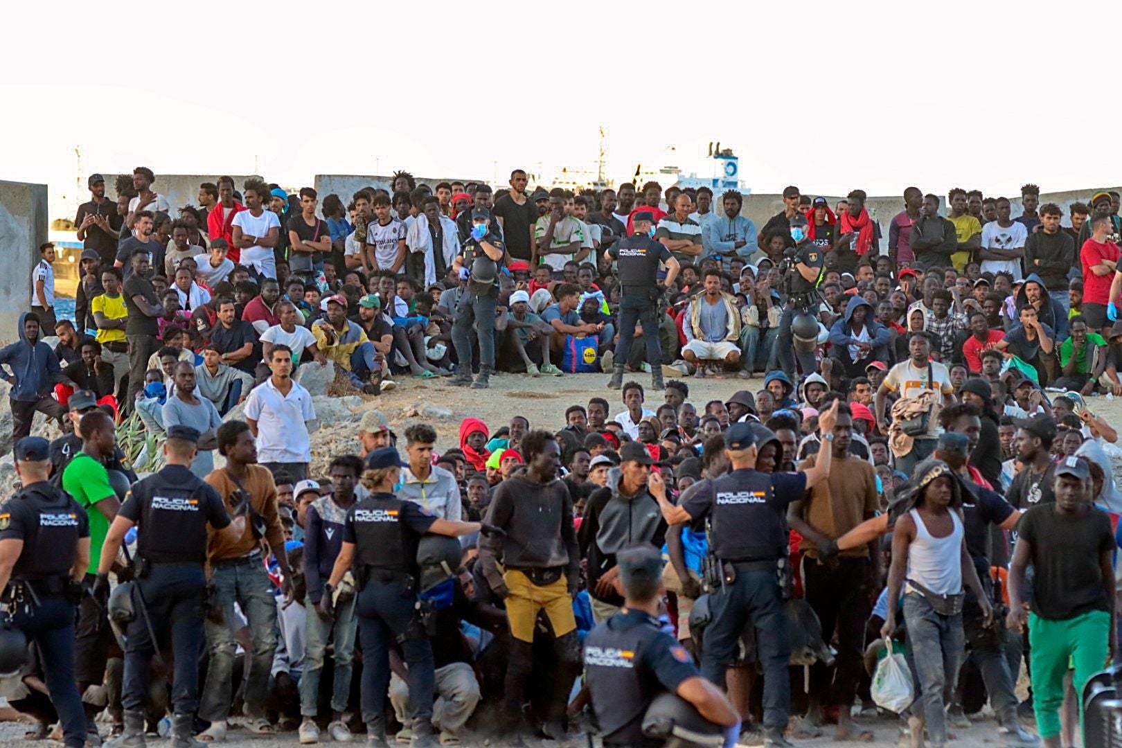 Viele Migranten sitzen und stehen an einem Strand, umgeben von zahlreichen Polizisten.