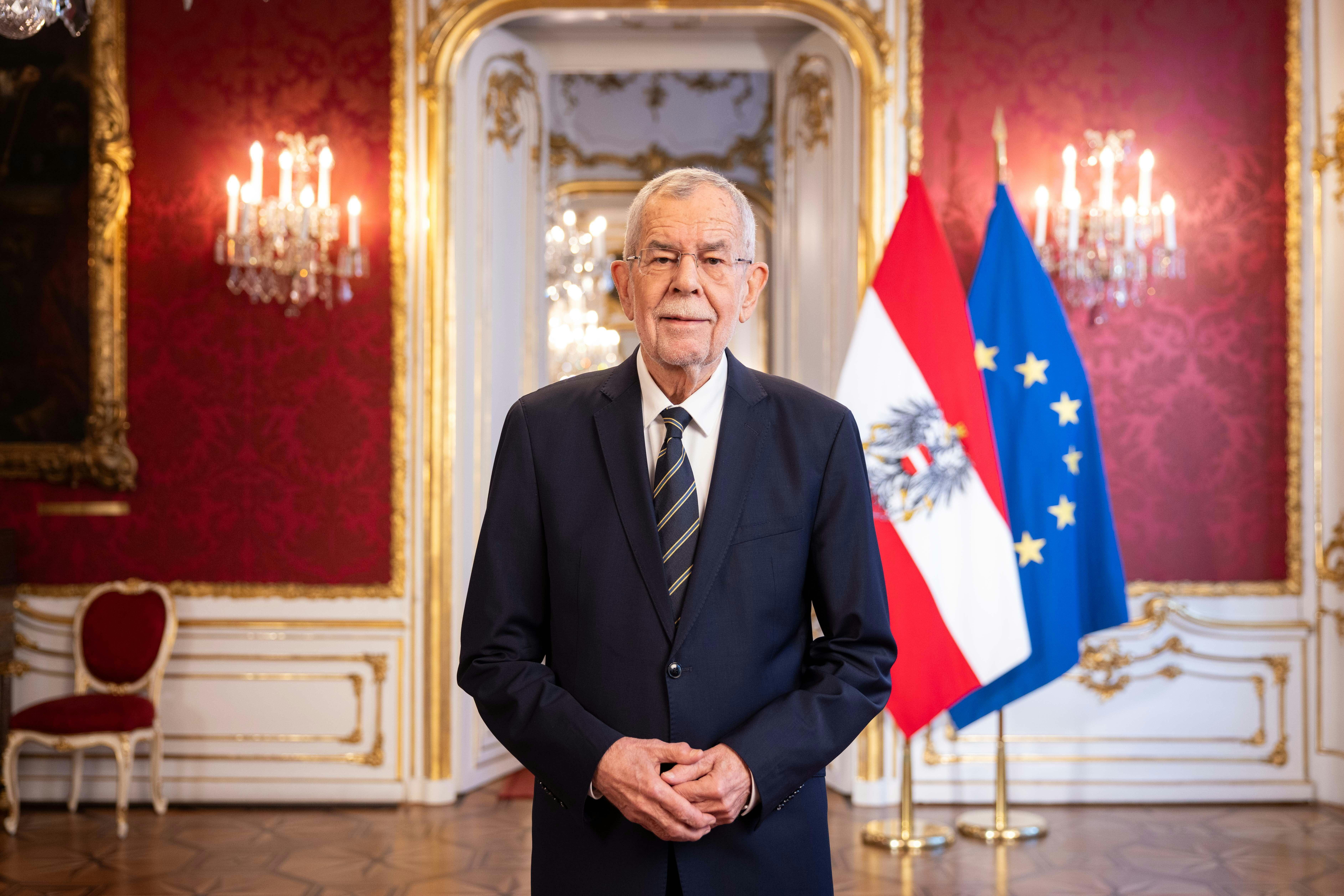 Ein älterer Mann im Anzug steht vor österreichischer und EU-Flagge in einem prunkvollen Saal.