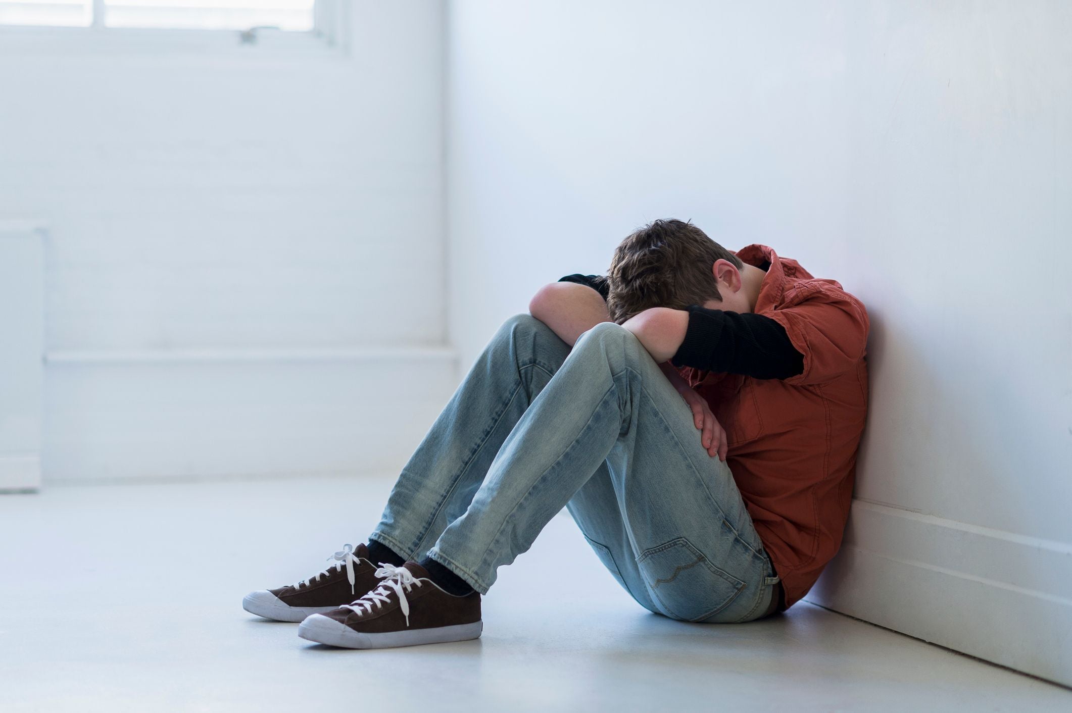 Jugendlicher sitzt traurig mit gesenktem Kopf an eine weiße Wand gelehnt auf dem Boden.