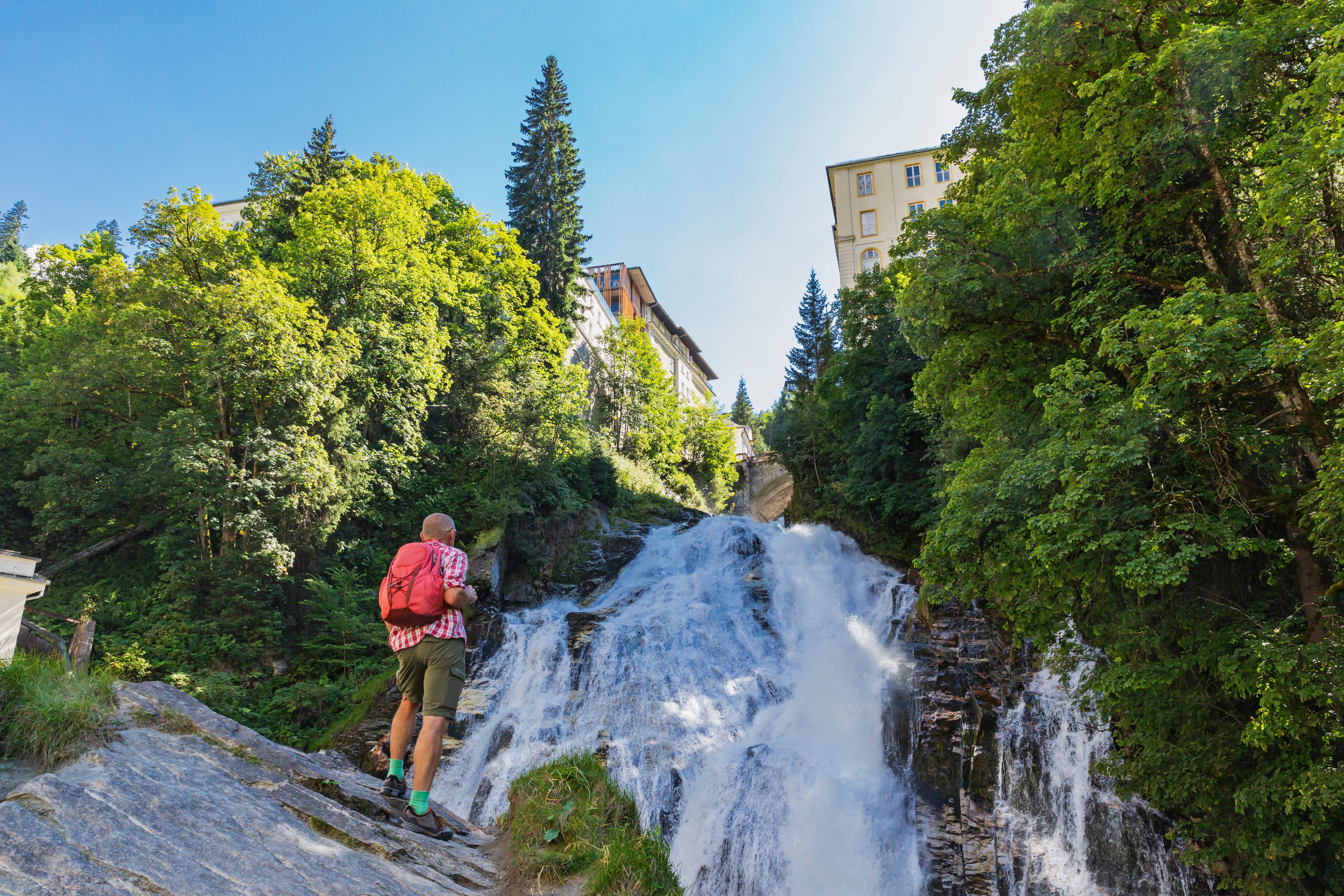 Ein Wanderer steht vor einem großen Wasserfall im Wald, im Hintergrund sind Gebäude am Hang.
