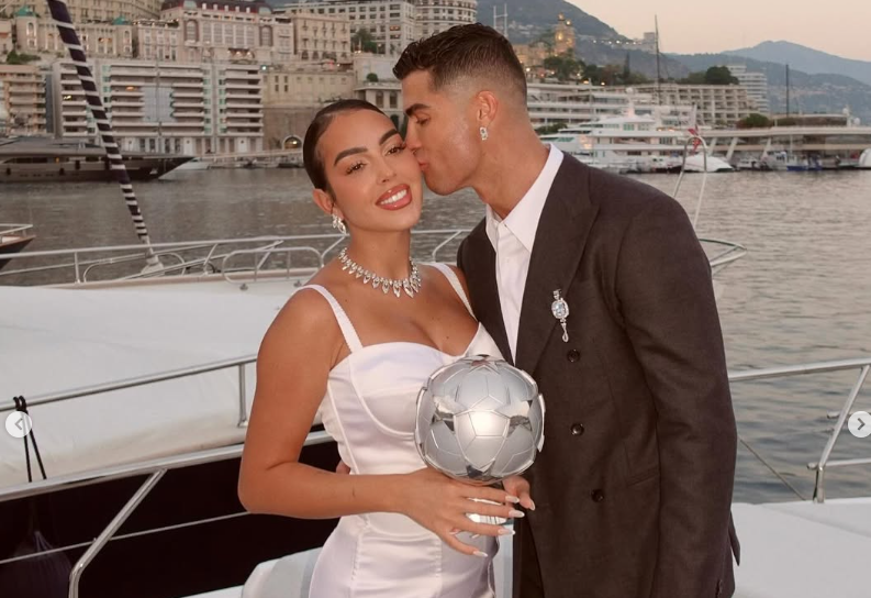Ein elegant gekleidetes Paar posiert auf einer Yacht, der Mann küsst die Frau auf die Stirn.