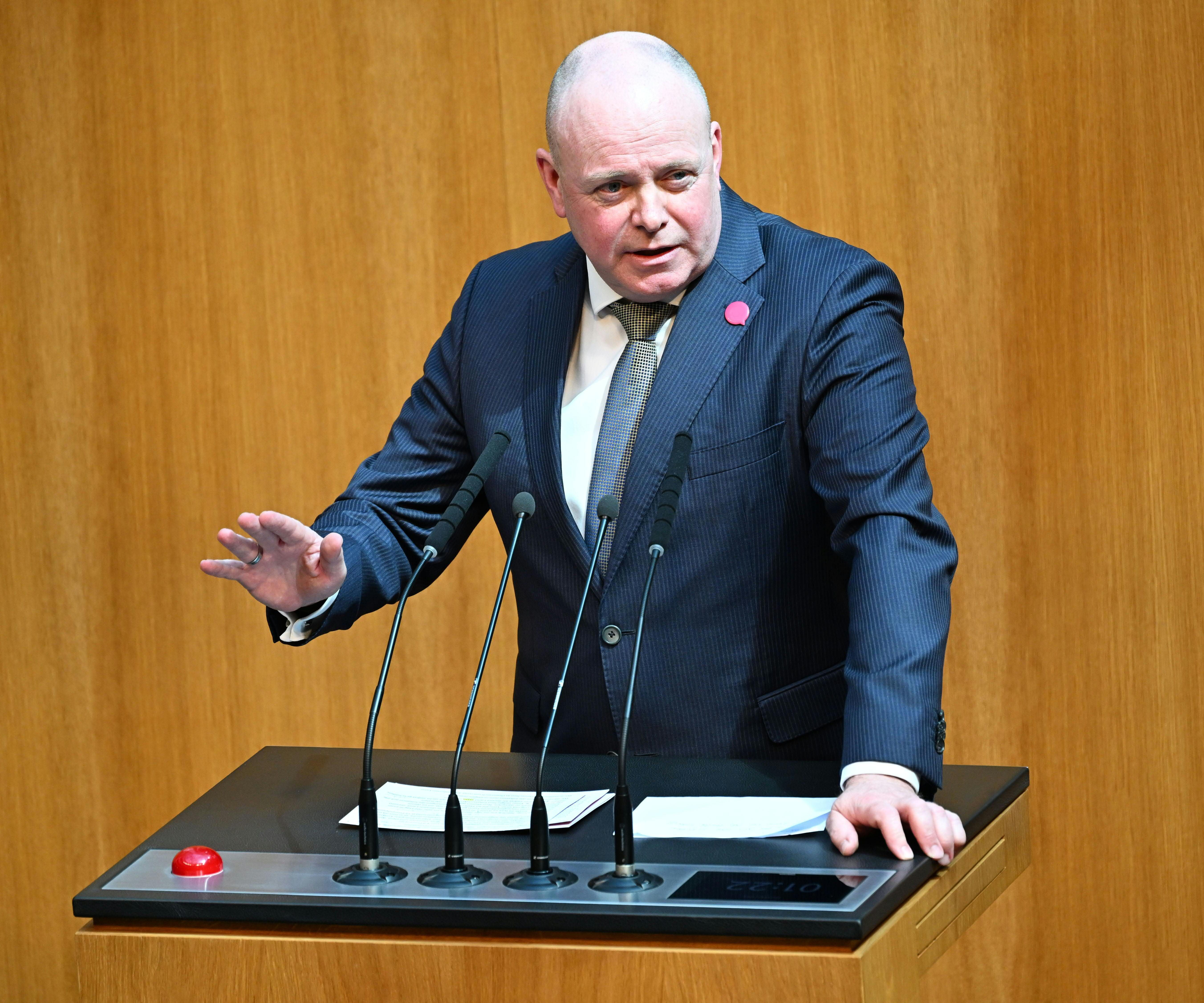 Ein Mann im Anzug spricht am Rednerpult im Parlament, vor ihm mehrere Mikrofone.