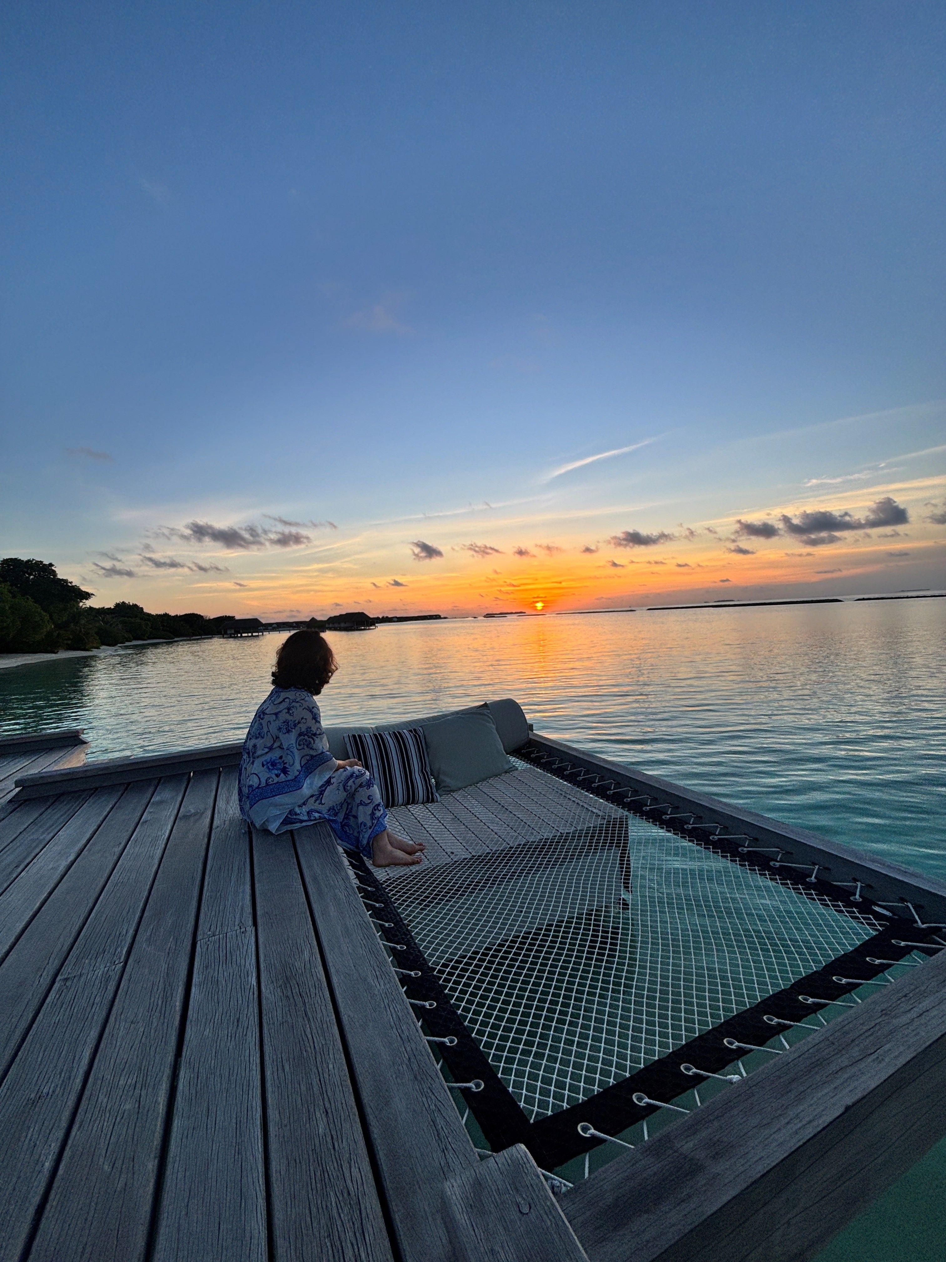 Person sitzt auf einem Holzdeck am Wasser und betrachtet den Sonnenuntergang auf Landaa Giravaru.