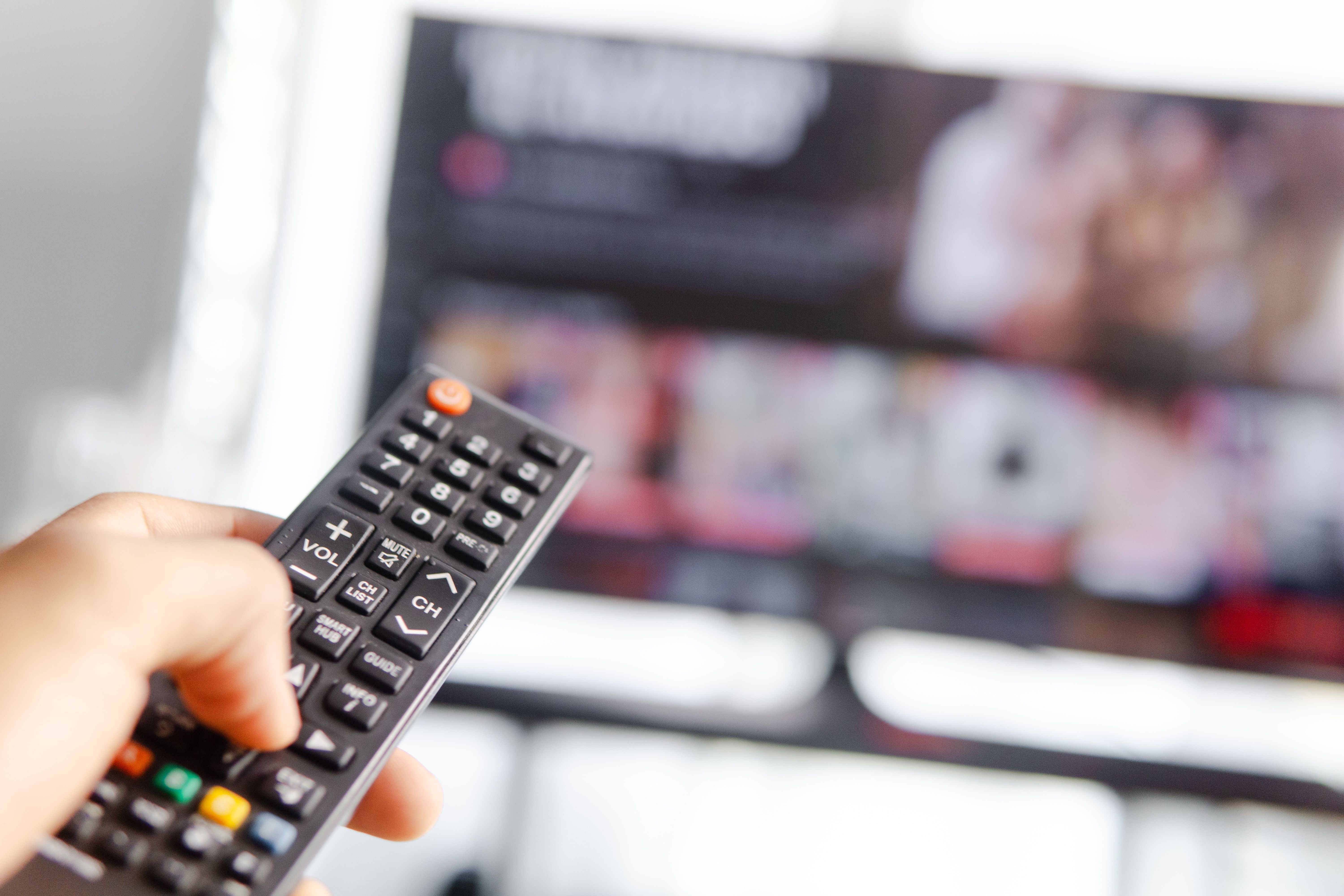 Hand hält eine Fernbedienung vor einem unscharfen Fernseher im Hintergrund.