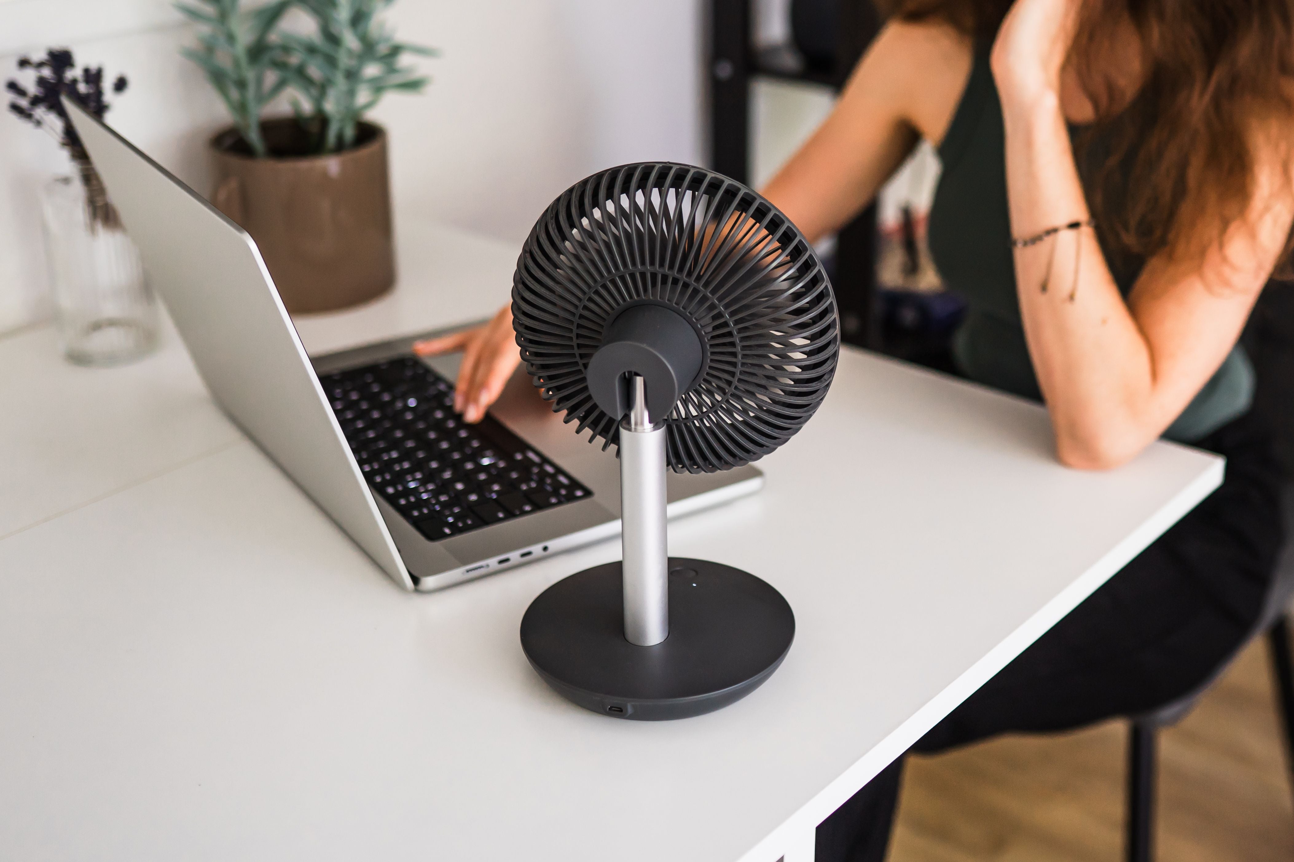 Kompakter schwarzer Ventilator auf Schreibtisch neben Laptop, Frau tippt im Hintergrund.