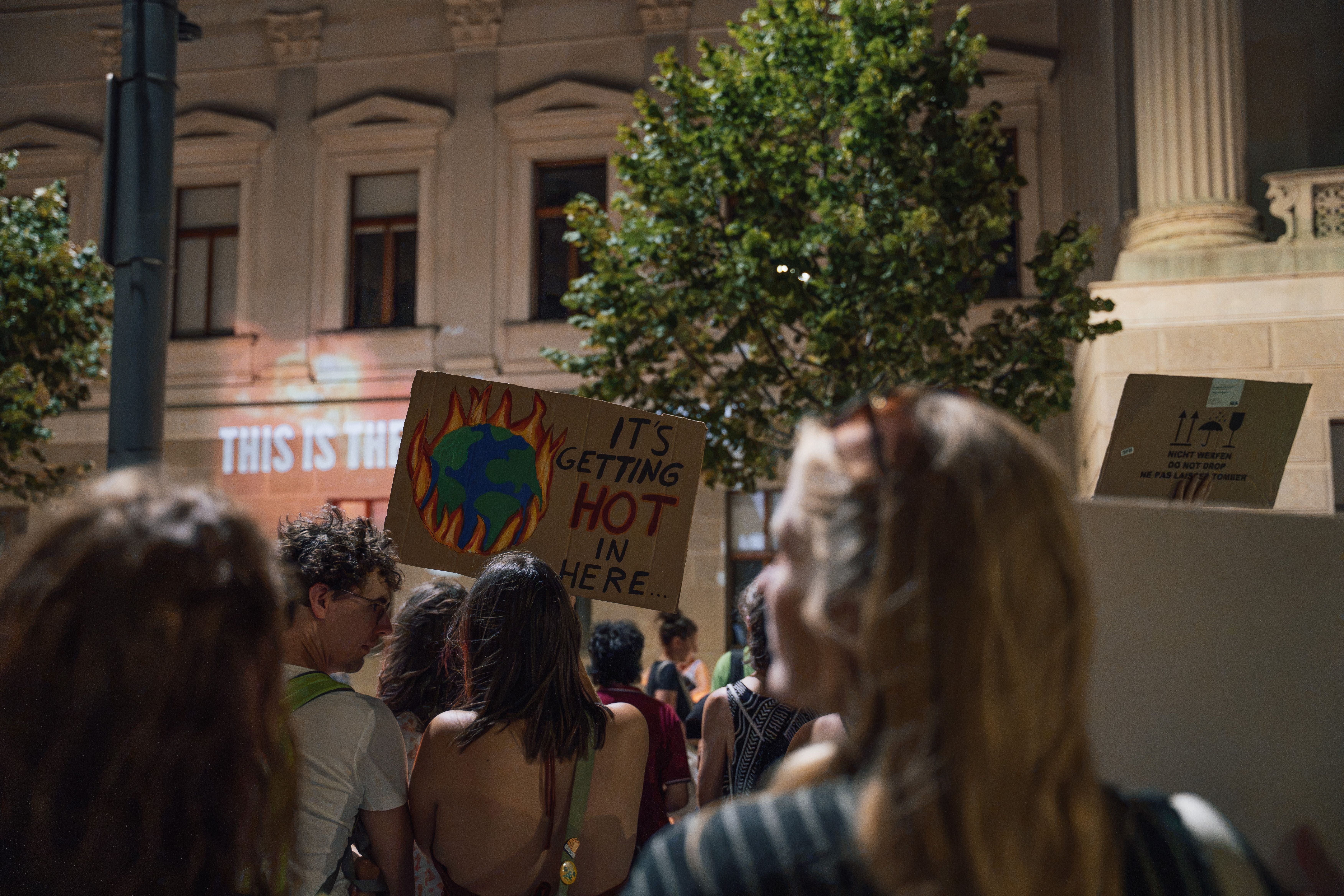 Menschen bei einer Hitze-Demo mit einem Schild: „It’s getting hot in here...“ und brennender Erde.