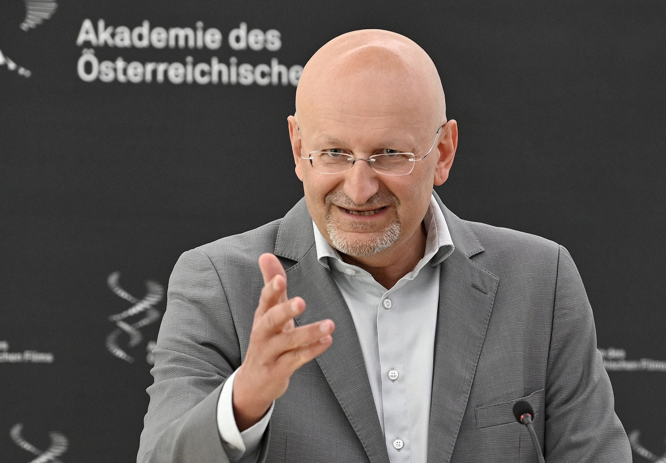Ein Mann im grauen Anzug spricht auf einer Pressekonferenz vor schwarzem Hintergrund.
