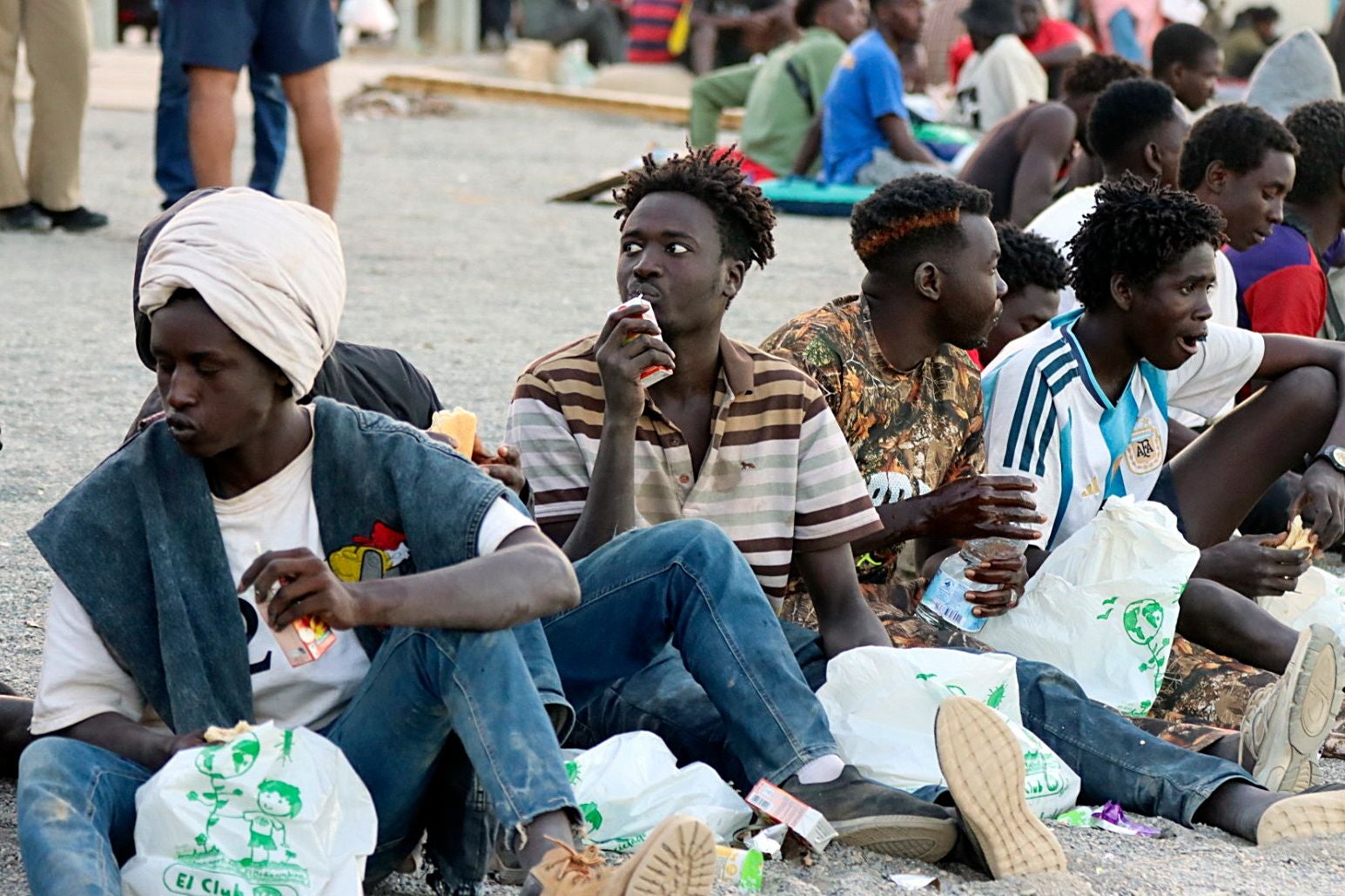 Mehrere Migranten sitzen im Freien und essen verteiltes Essen aus Plastiktüten.