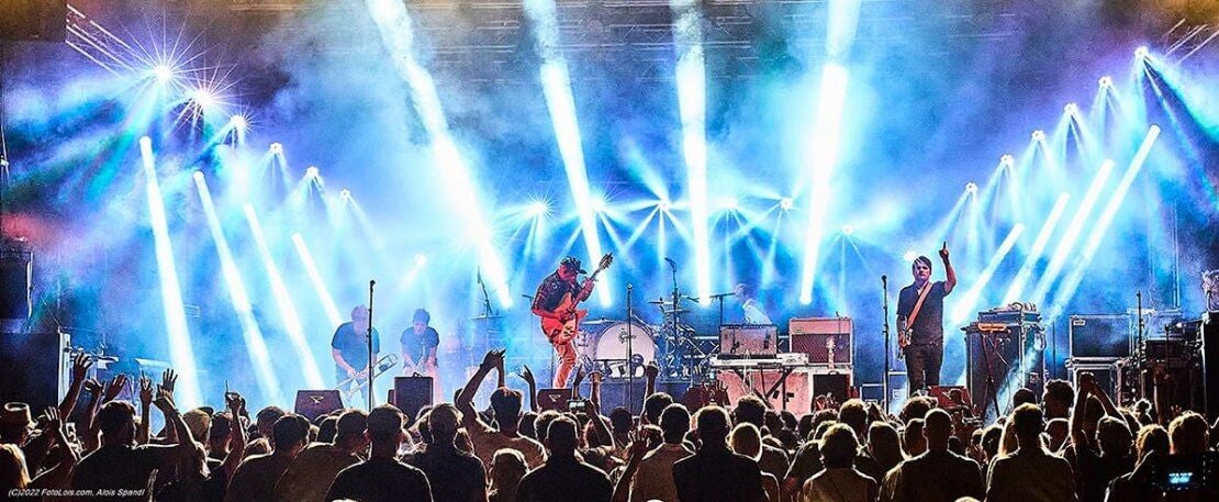 Eine Band spielt auf einer hell beleuchteten Bühne vor jubelndem Publikum bei einem Konzert.