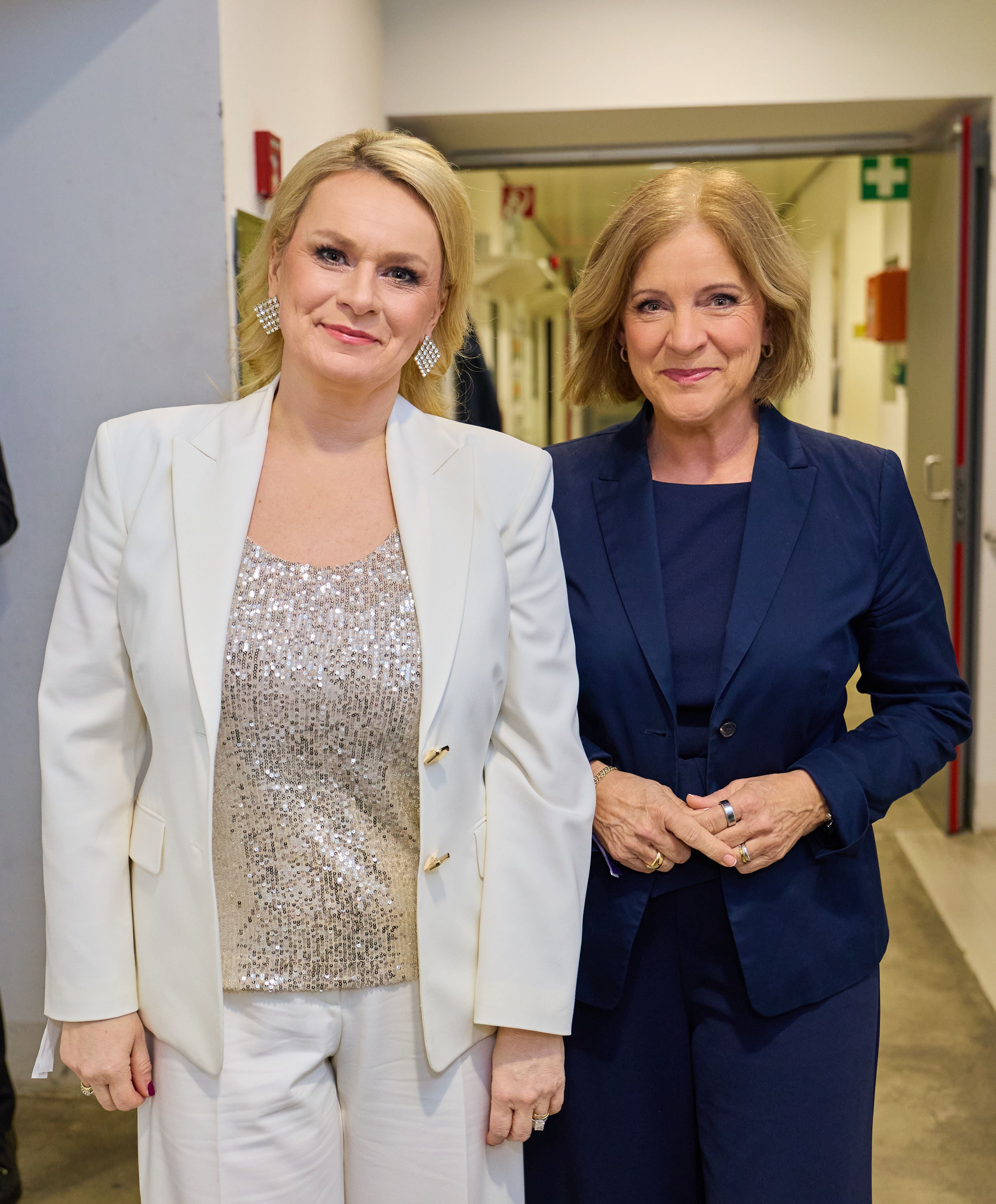 Zwei Frauen in eleganten Hosenanzügen stehen nebeneinander und lächeln in die Kamera.