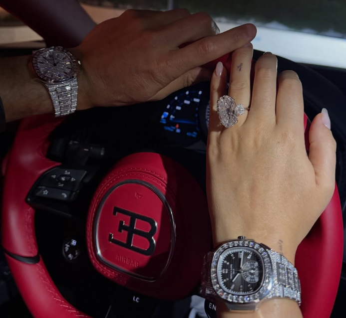 Zwei Hände mit Diamantuhren und großem Ring auf einem roten Bugatti-Lenkrad.