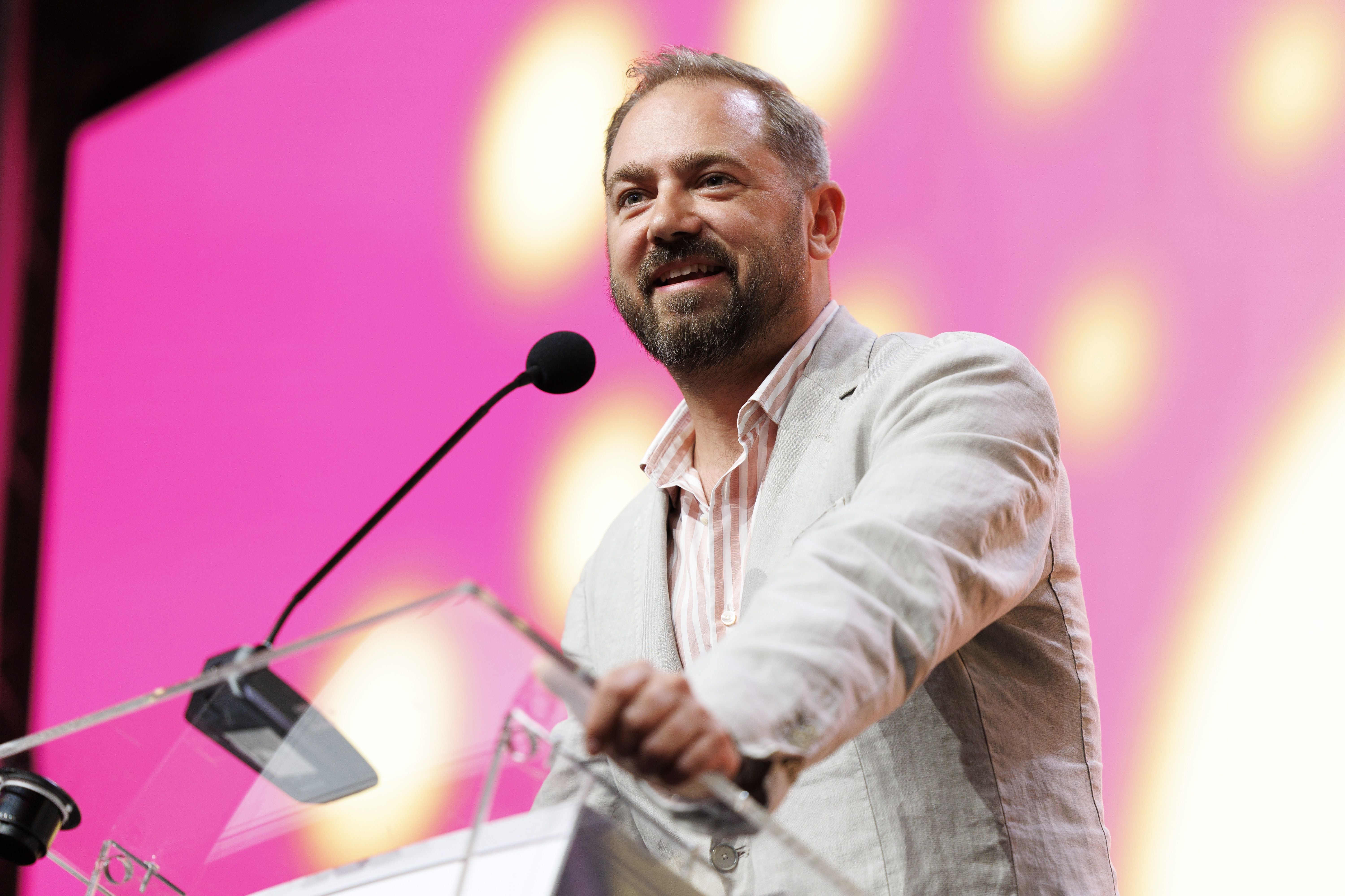 Ein Mann spricht lächelnd am Rednerpult vor einem pinken Hintergrund mit Mikrofon.