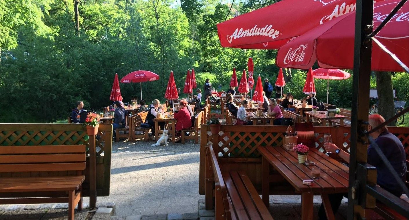 Gäste sitzen an Holztischen im sonnigen Biergarten mit roten Sonnenschirmen und Bäumen im Hintergrund.