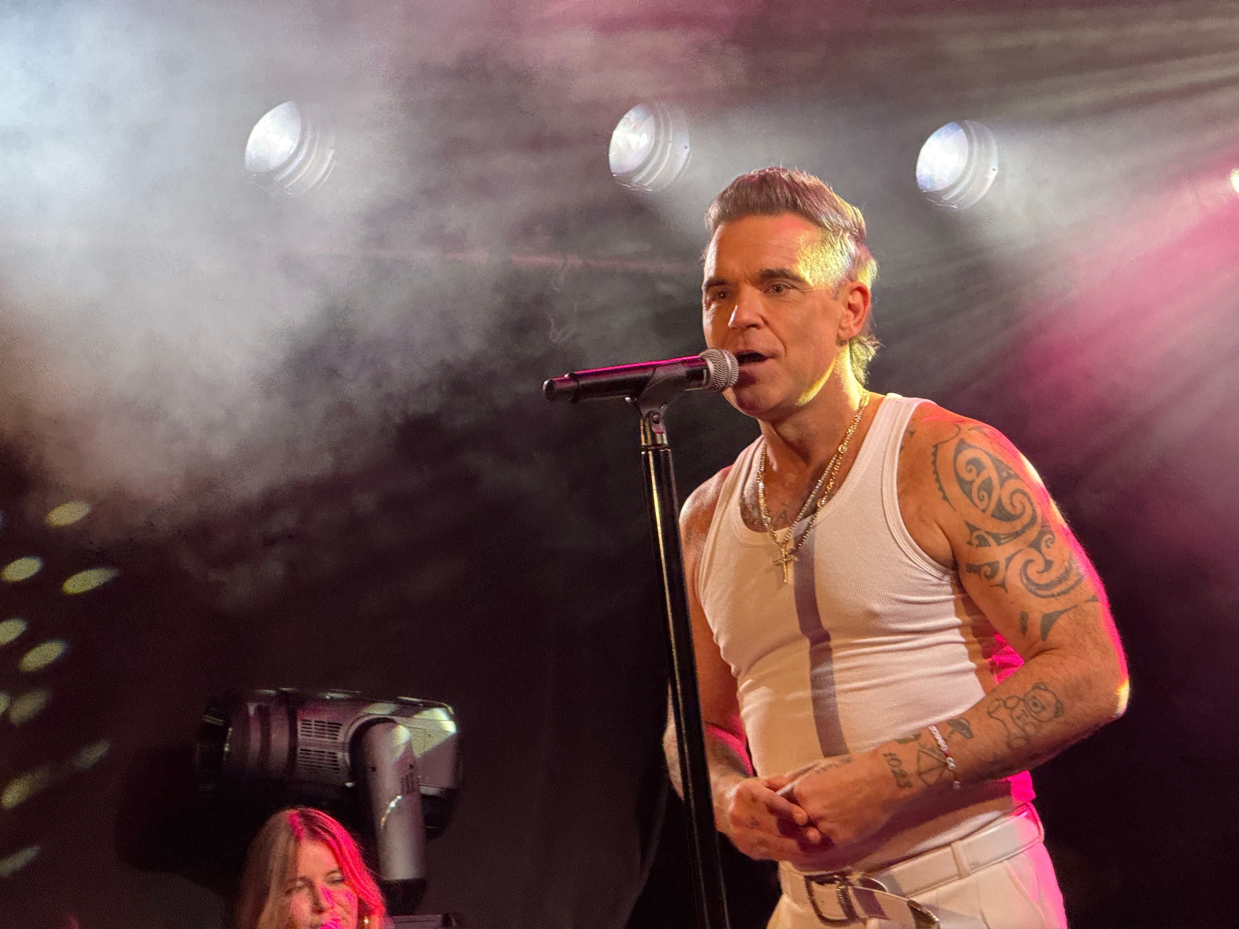 Mann mit Tattoos singt auf einer Bühne im Nebel in weißem Unterhemd und Hose.