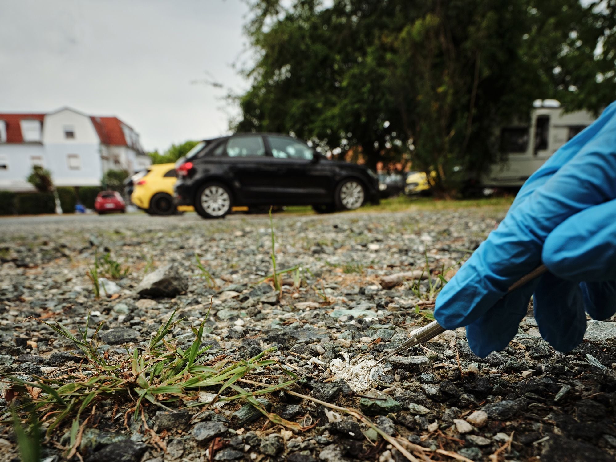 Hand mit blauem Handschuh nimmt mit Pinzette eine Probe von mit Asbest verseuchtem Kies auf einem Parkplatz.
